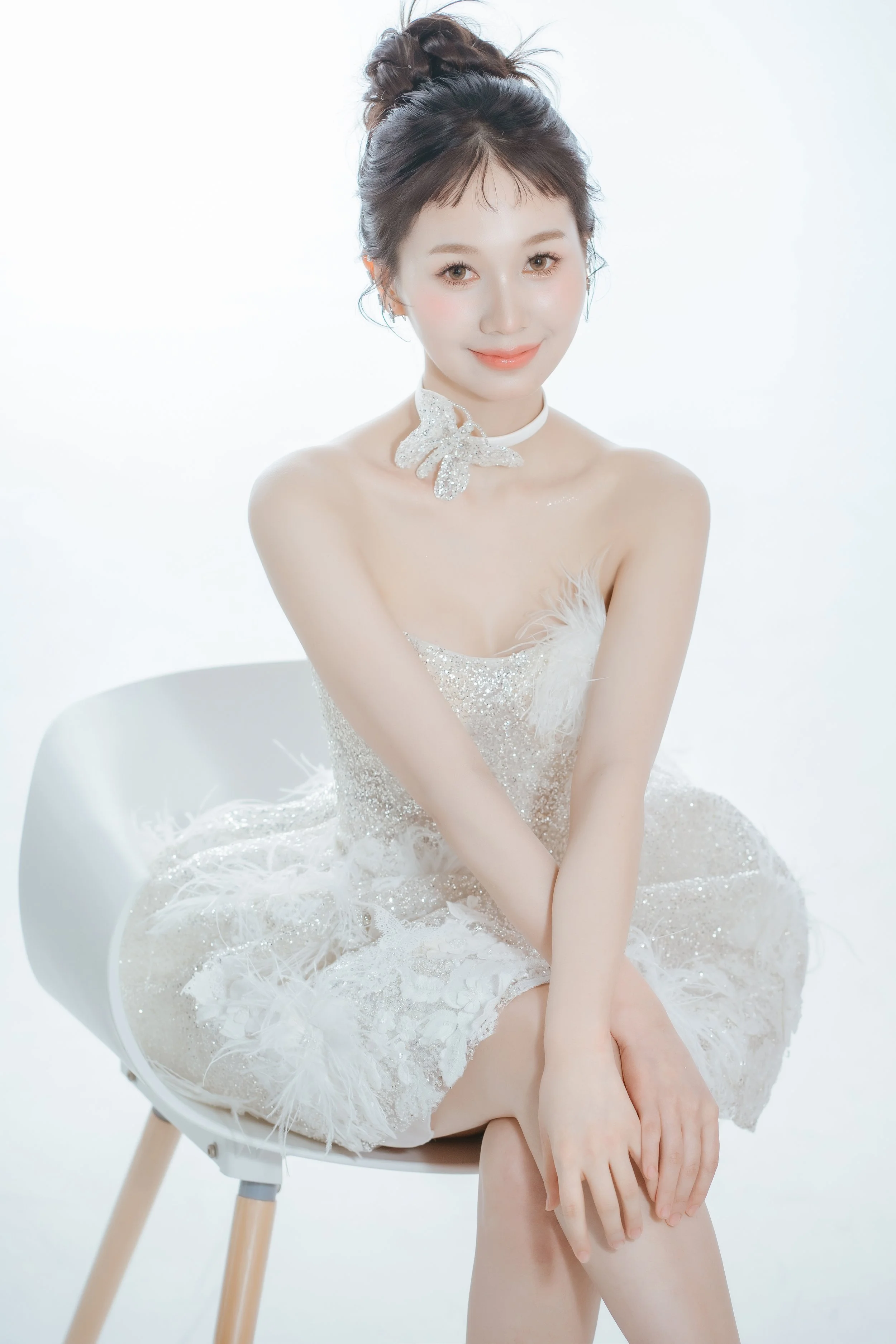 Young woman with dark hair styled in an updo, sitting on a white chair, wearing a white dress with lace details and a butterfly-shaped necklace, smiling softly at the camera, against a plain white background.