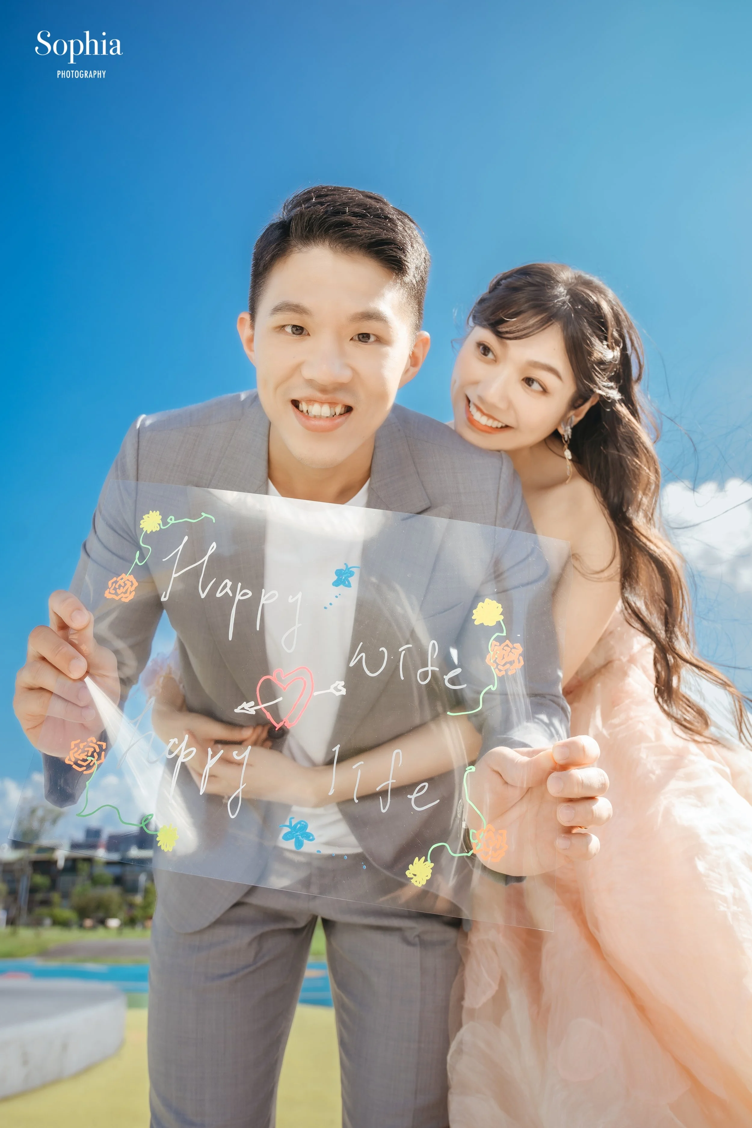 A happy couple outdoors under a clear blue sky, holding a transparent sign with colorful flowers and the words 'Happy Wife Happy Life' written on it, smiling at each other.