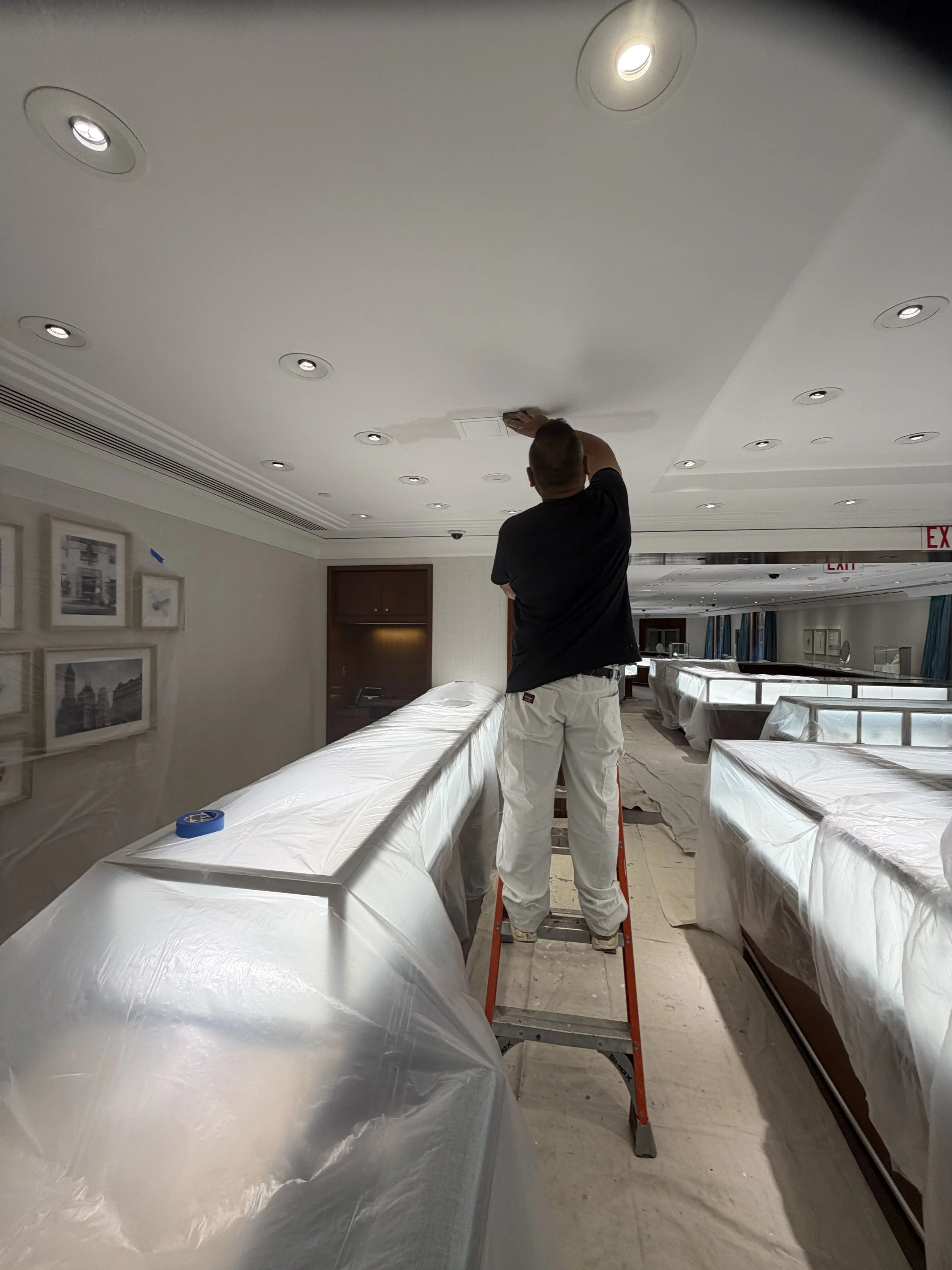 A person standing on a ladder cleaning or repairing a ceiling with multiple recessed lights, in a room that appears to be under renovation or maintenance, with furniture covered in protective plastic.