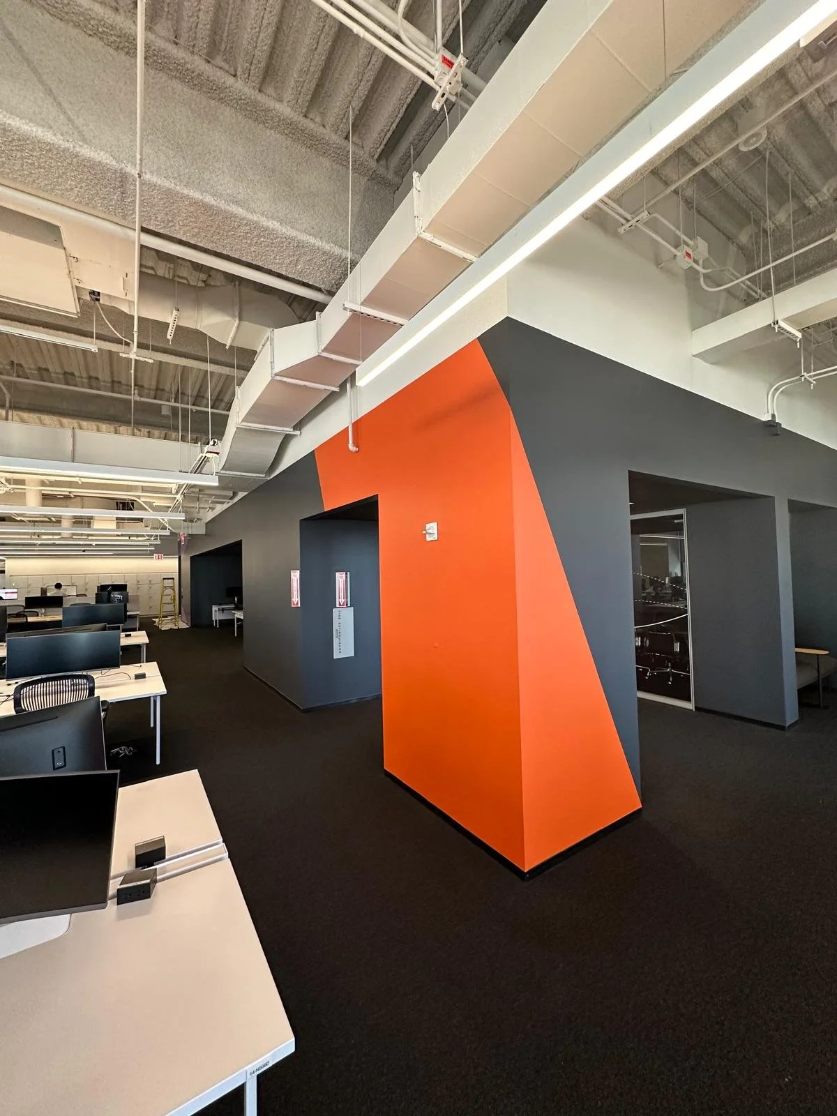 Interior of an office with black carpet, white desks, and black chairs. A prominent wall features a geometric orange and gray design. Exposed ceiling with white ducts and lighting fixtures.