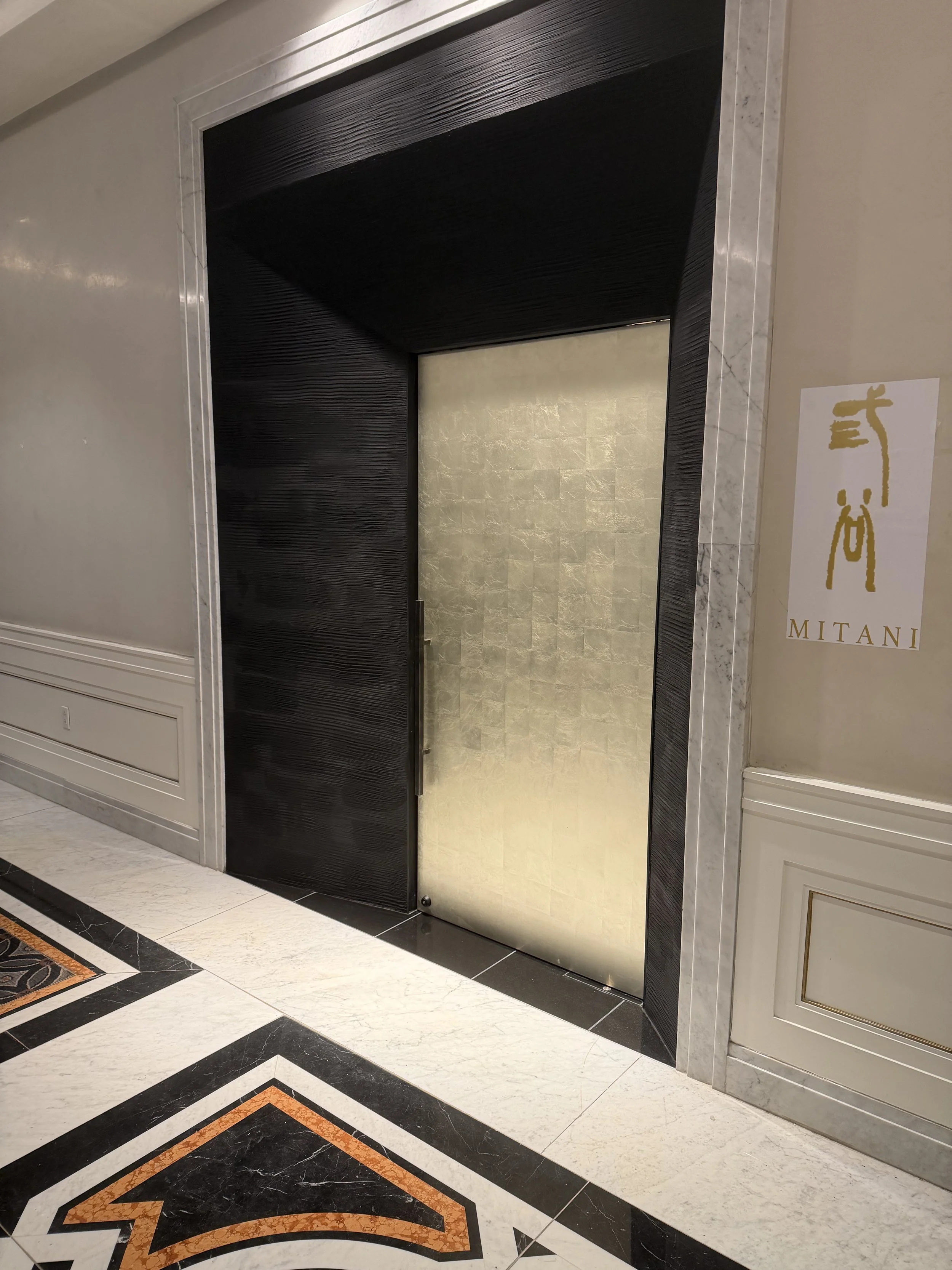 An elevator with gold-colored doors in a luxurious hotel lobby, with an ornate marble floor and decorative wall paneling around the elevator.