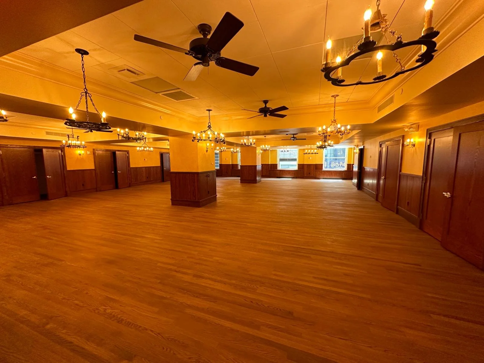 An empty banquet or event hall with warm yellow walls, wooden accents, multiple chandeliers, and ceiling fans, illuminated by wall-mounted sconces and chandeliers, with several closed doors along the walls and large windows at the far end.