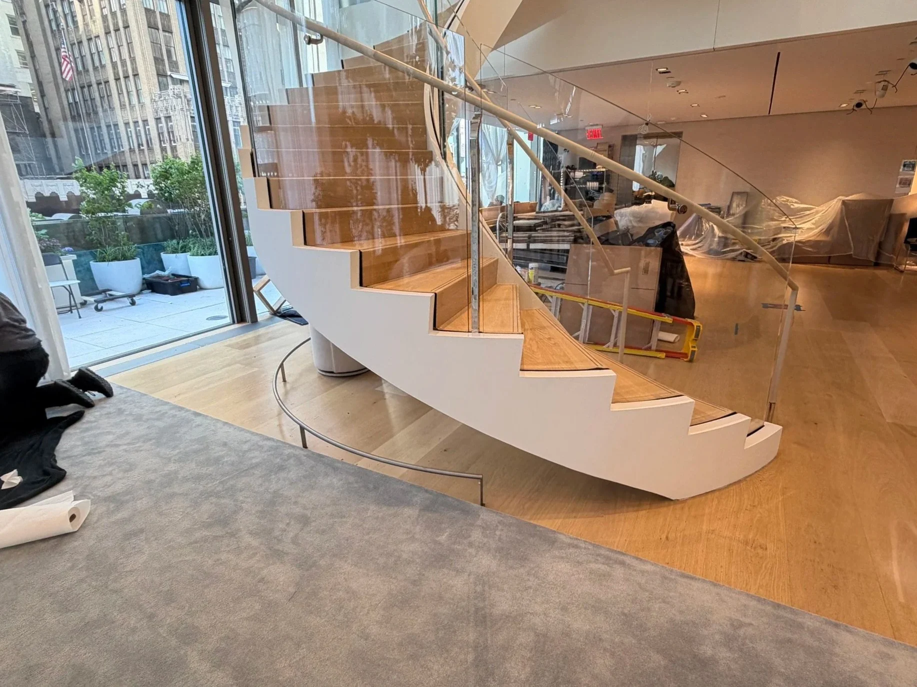 Interior space with a curved staircase featuring wooden steps and glass railings, and large windows revealing buildings and outdoor plants outside.