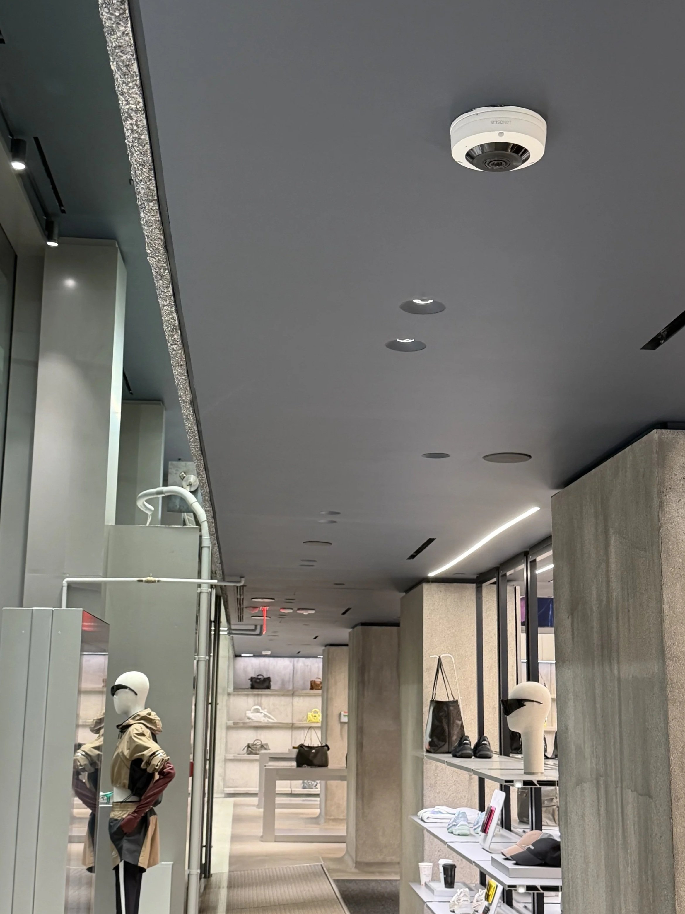 Interior of a modern retail store with a gray ceiling, display shelves with hats, drinks, and accessories, and mannequins wearing helmets and casual clothing, with bright lighting and a contemporary design.