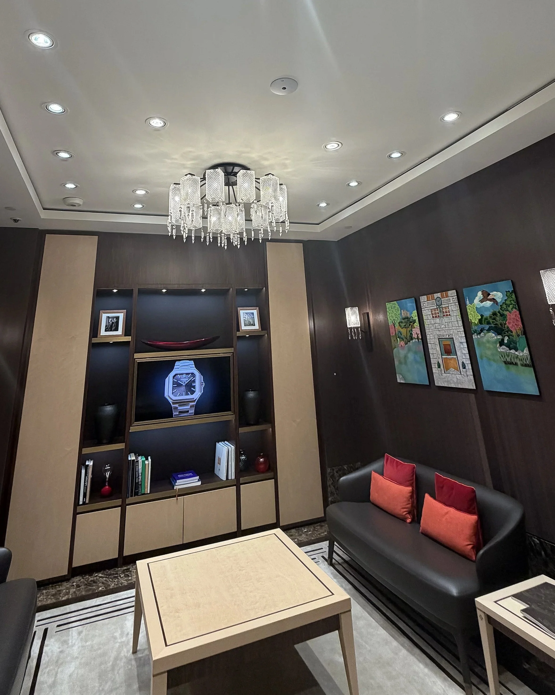 Interior view of a modern living room with a black sofa adorned with red and orange pillows, a beige coffee table, a wooden bookshelf with decorative items and framed photos, a chandelier hanging from the ceiling, and colorful artworks on the dark wa