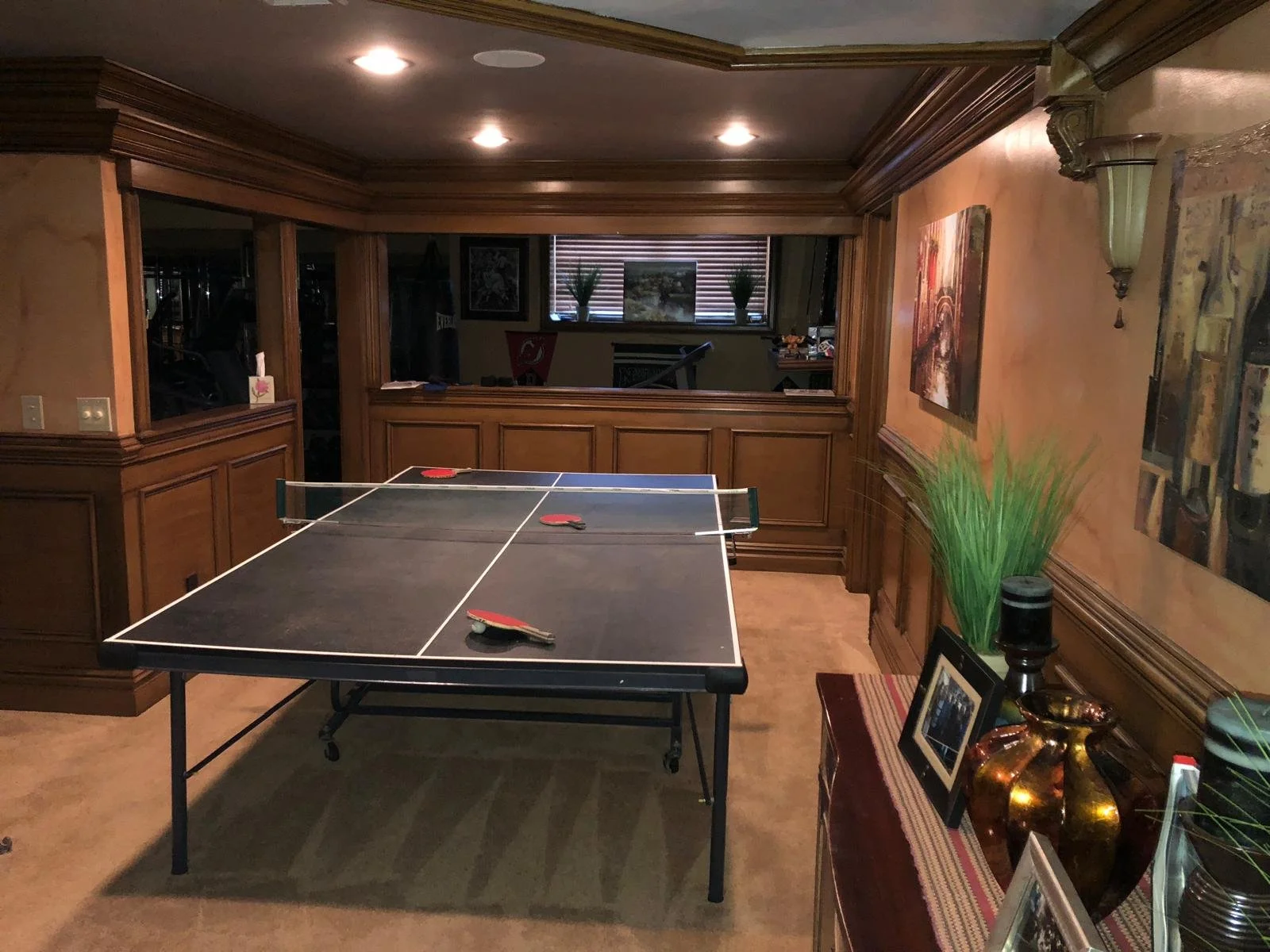 A recreational room with a ping pong table, wooden wall paneling, artwork on the walls, a window with blinds, and decorative items including vases and framed photos.