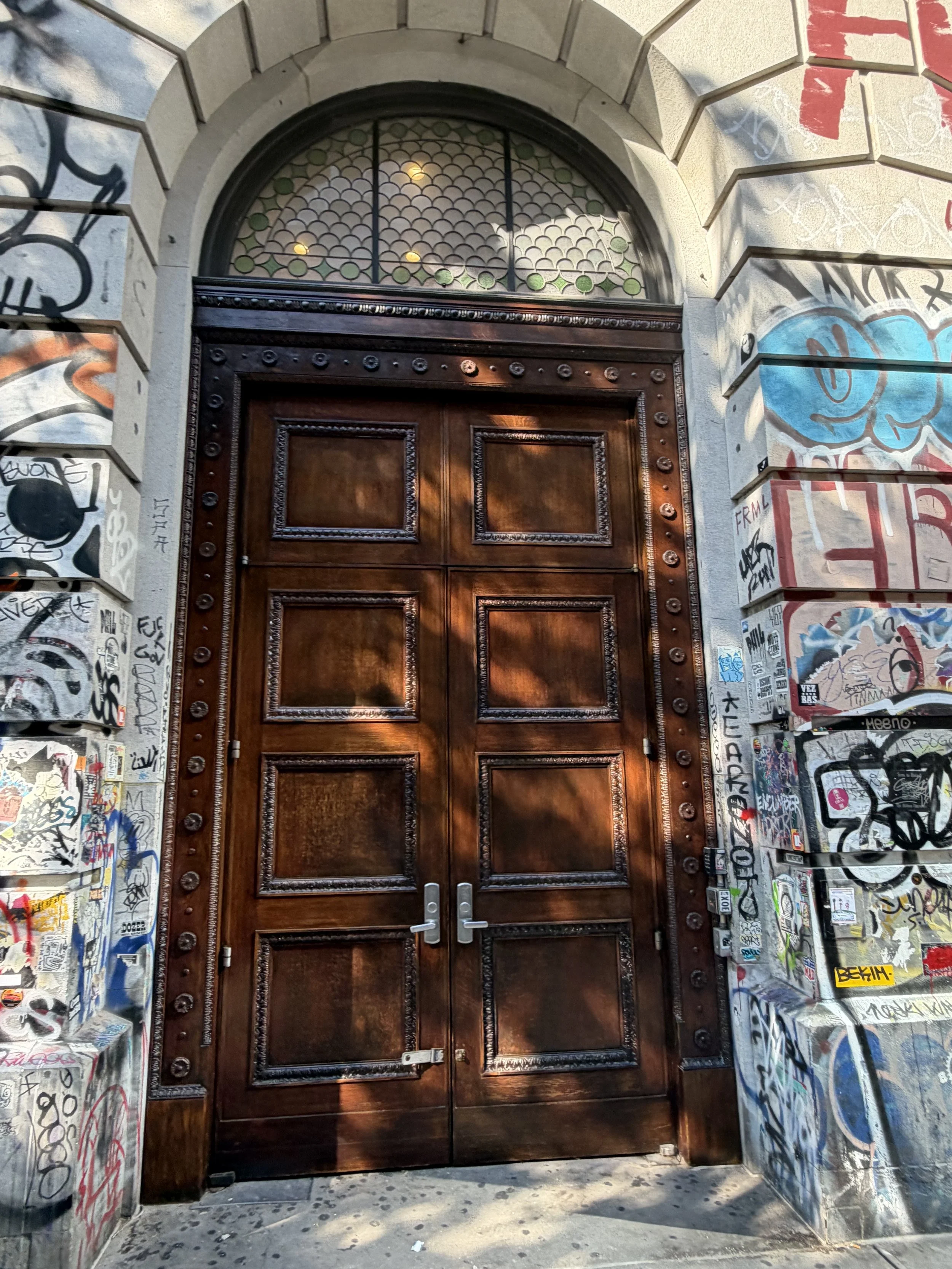 A large wooden door set in a stone archway, with stained glass window above, covered in colorful graffiti on the surrounding walls and metal panels.