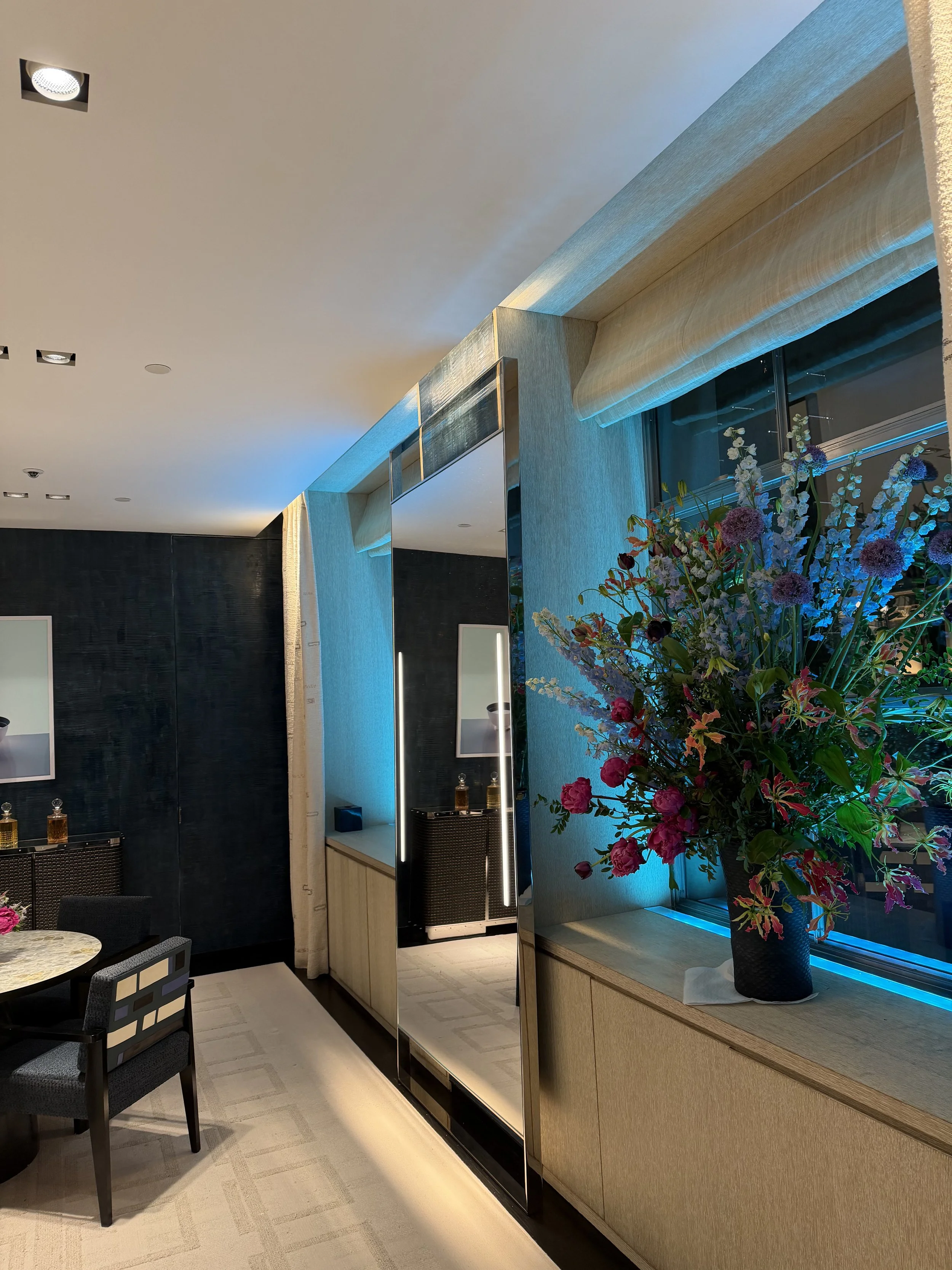 Interior of a modern room with a beige carpet, a large floral arrangement in a black vase, a light-colored wooden cabinet, and a mirror with vertical lights, illuminated by both natural and artificial lighting.