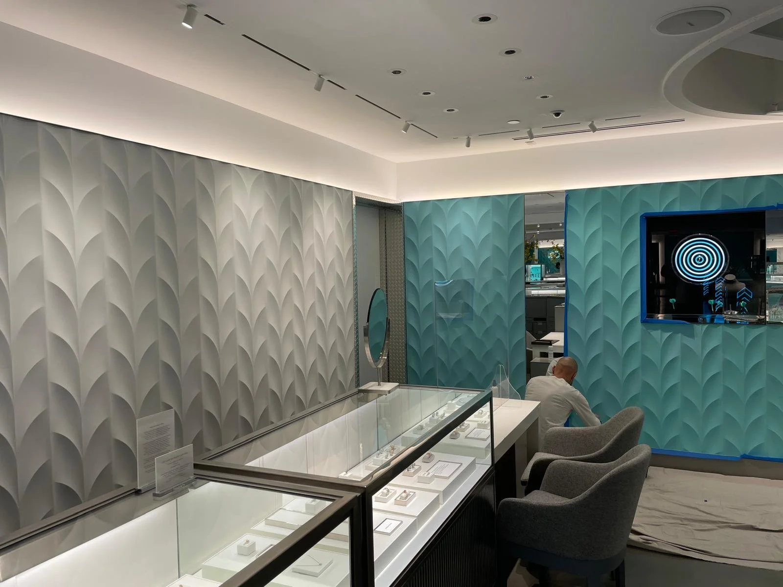 Interior of a modern showroom or gallery with textured white and blue walls, display cases with jewelry, and a person sitting on a chair.
