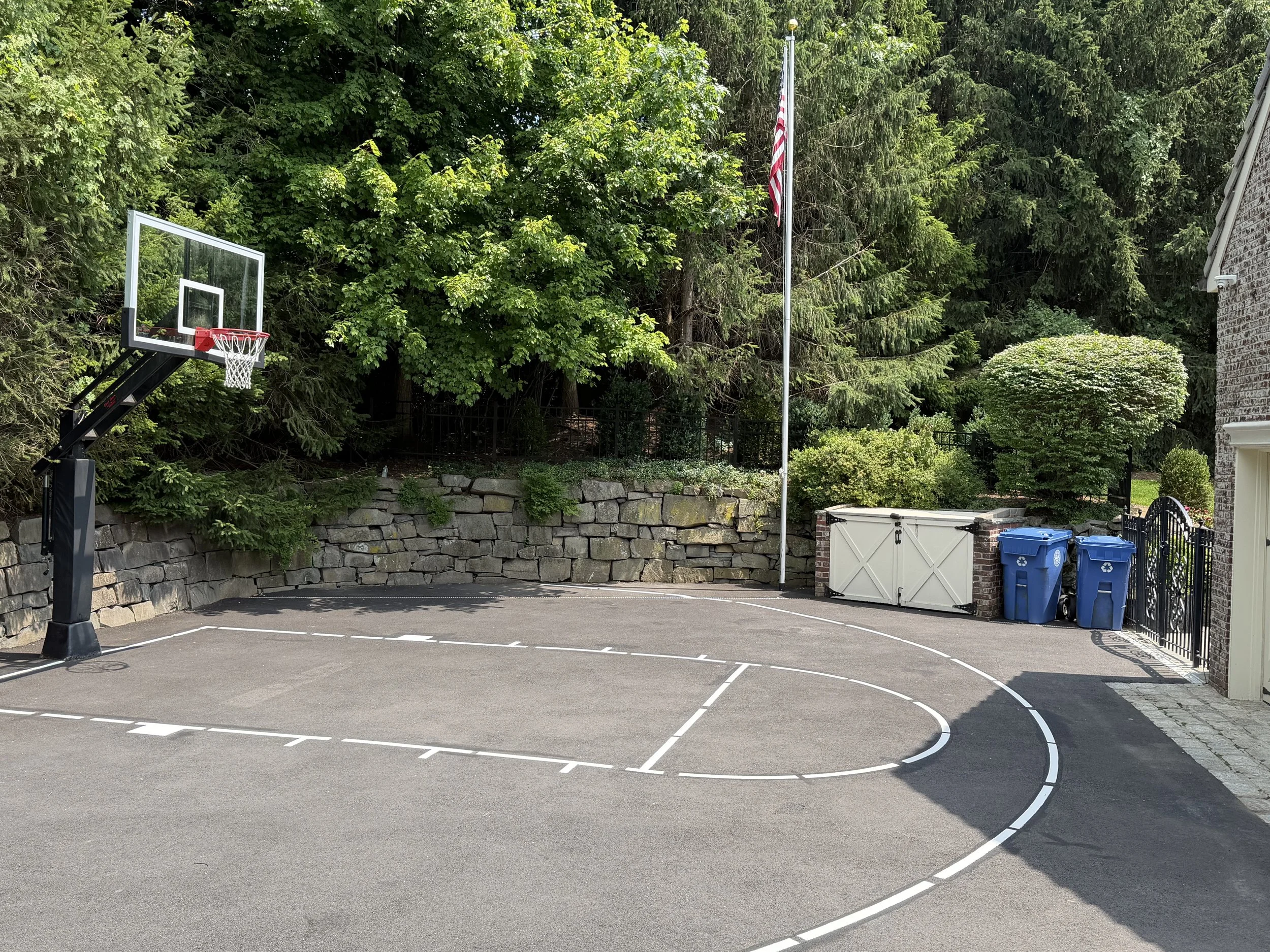 A residential backyard basketball court with a basketball hoop on the left, surrounded by green trees and shrubs. There are recycling bins and a white shed on the right, and a stone retaining wall in the background.