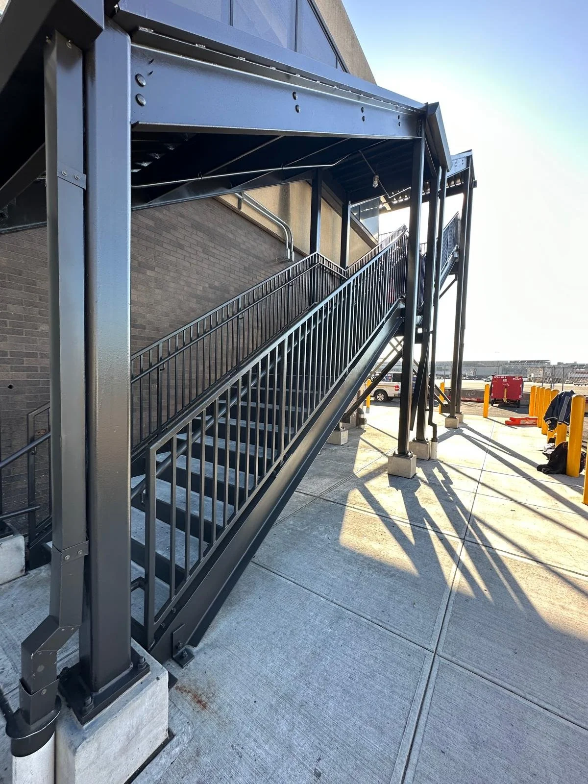 Exterior metal staircase with handrails leading to a higher level, set against a brick building wall, with parked cars and yellow safety posts in the background, and sunlight casting shadows on the concrete ground.