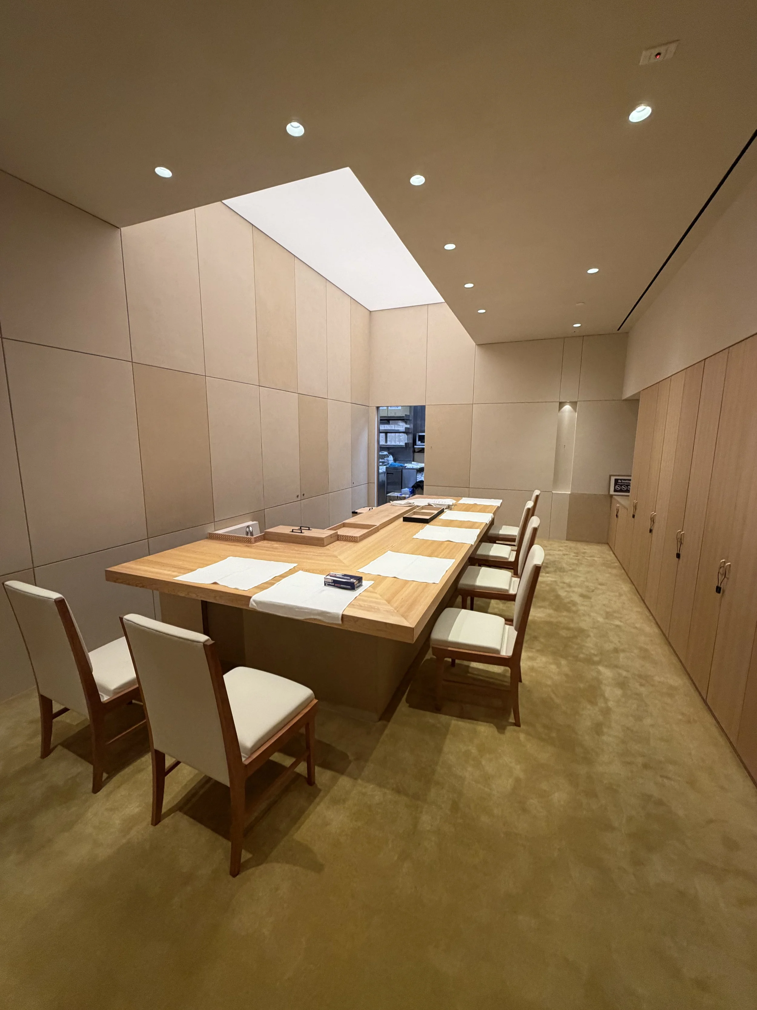 A modern dining area with a rectangular wooden table surrounded by eight white chairs, set with white placemats and small wooden trays, in a room with beige walls and carpet, ceiling lights, and a small window view of a kitchen or working area outside.