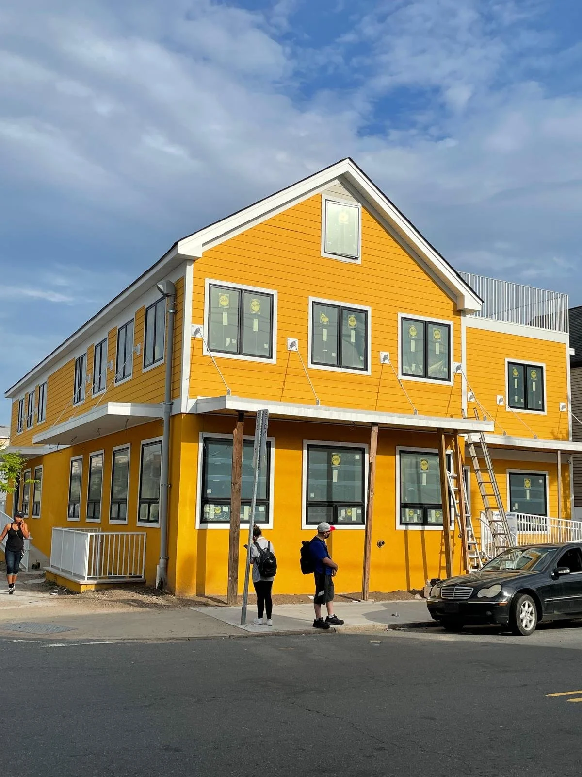 Yellow multi-story building under construction with black-framed windows, a ladder on the side, and pedestrians nearby on a street. A dark car is parked in front.
