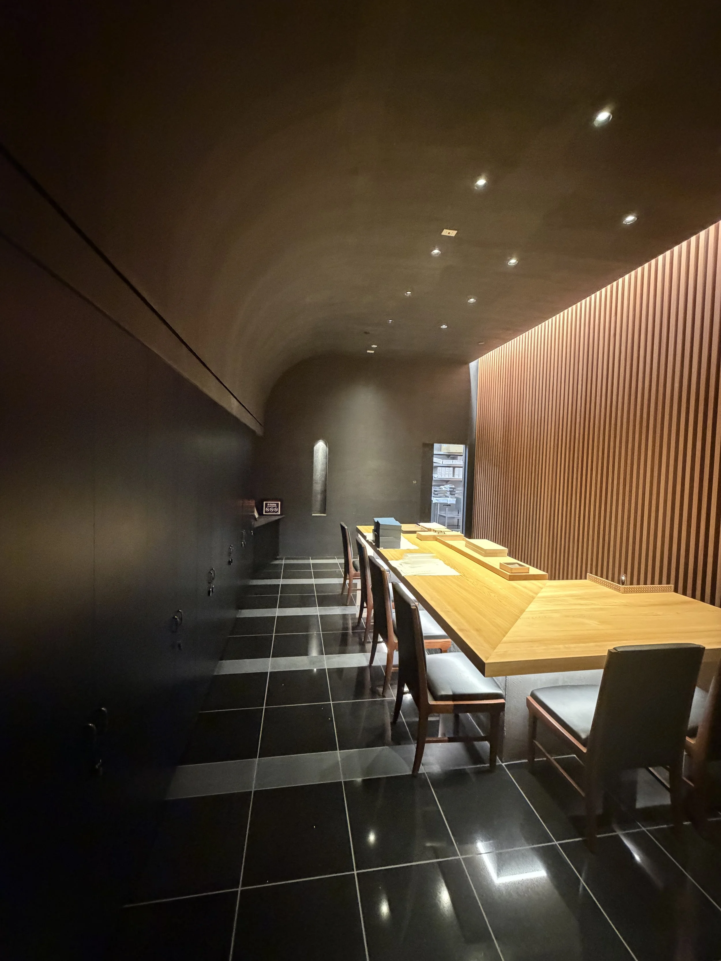 A modern interior with a dark ceiling and walls, a wooden table with chairs, and a wall with vertical wooden slats. The space is dimly lit with small ceiling lights and lights on the wall.
