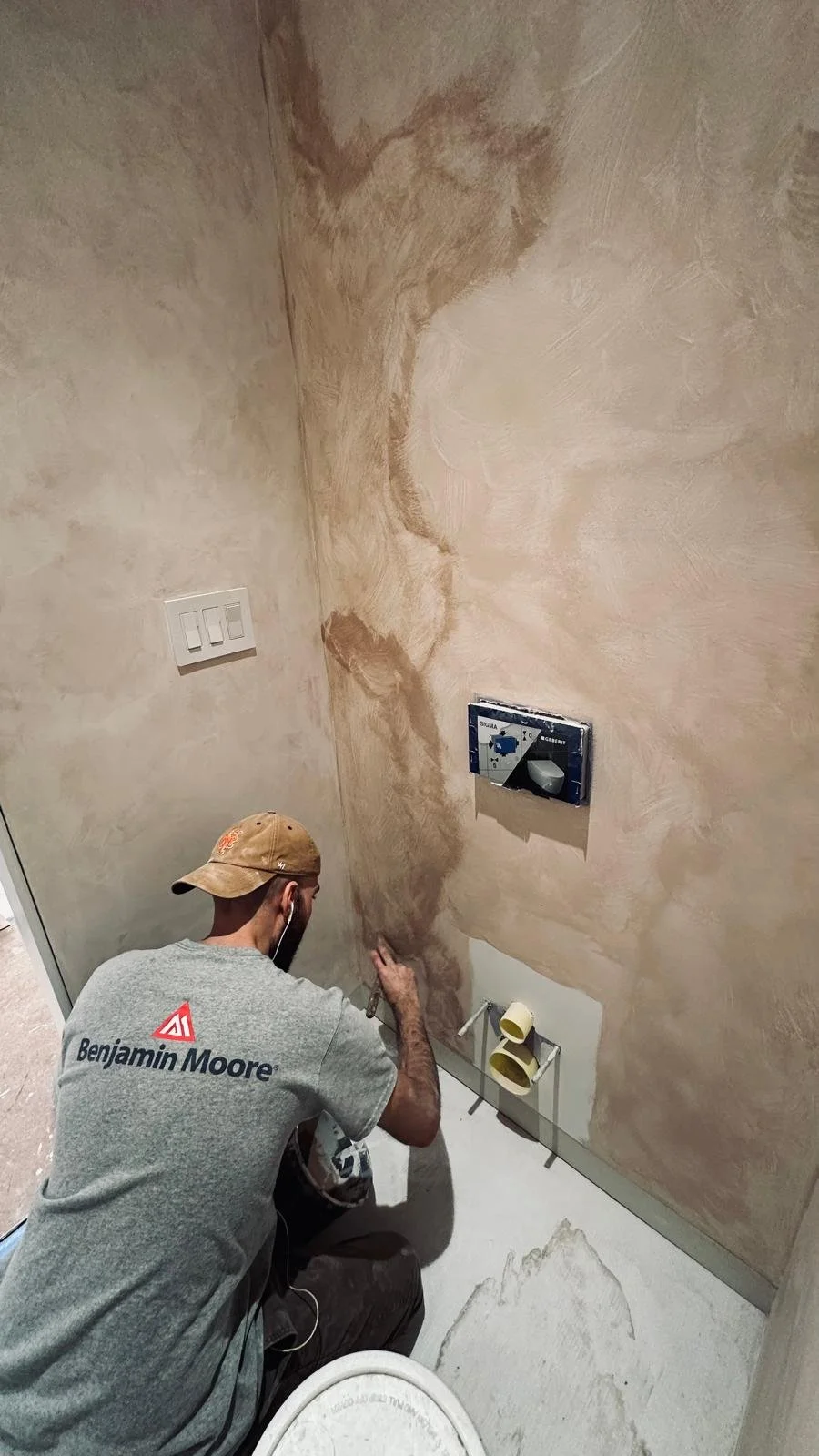 A man wearing a gray Benjamin Moore t-shirt and a tan cap is working on a bathroom wall that has a faux finish in shades of brown and beige. He is kneeling and appears to be painting or applying a finishing touch near the lower part of the wall, which has plumbing fixtures for a toilet.