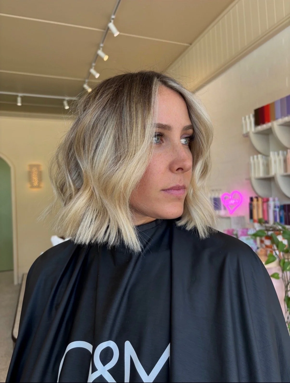 A woman with shoulder-length blonde hair styled in loose waves, wearing a black salon cape, standing inside a salon with shelves of hair products and a neon pink heart sign in the background.