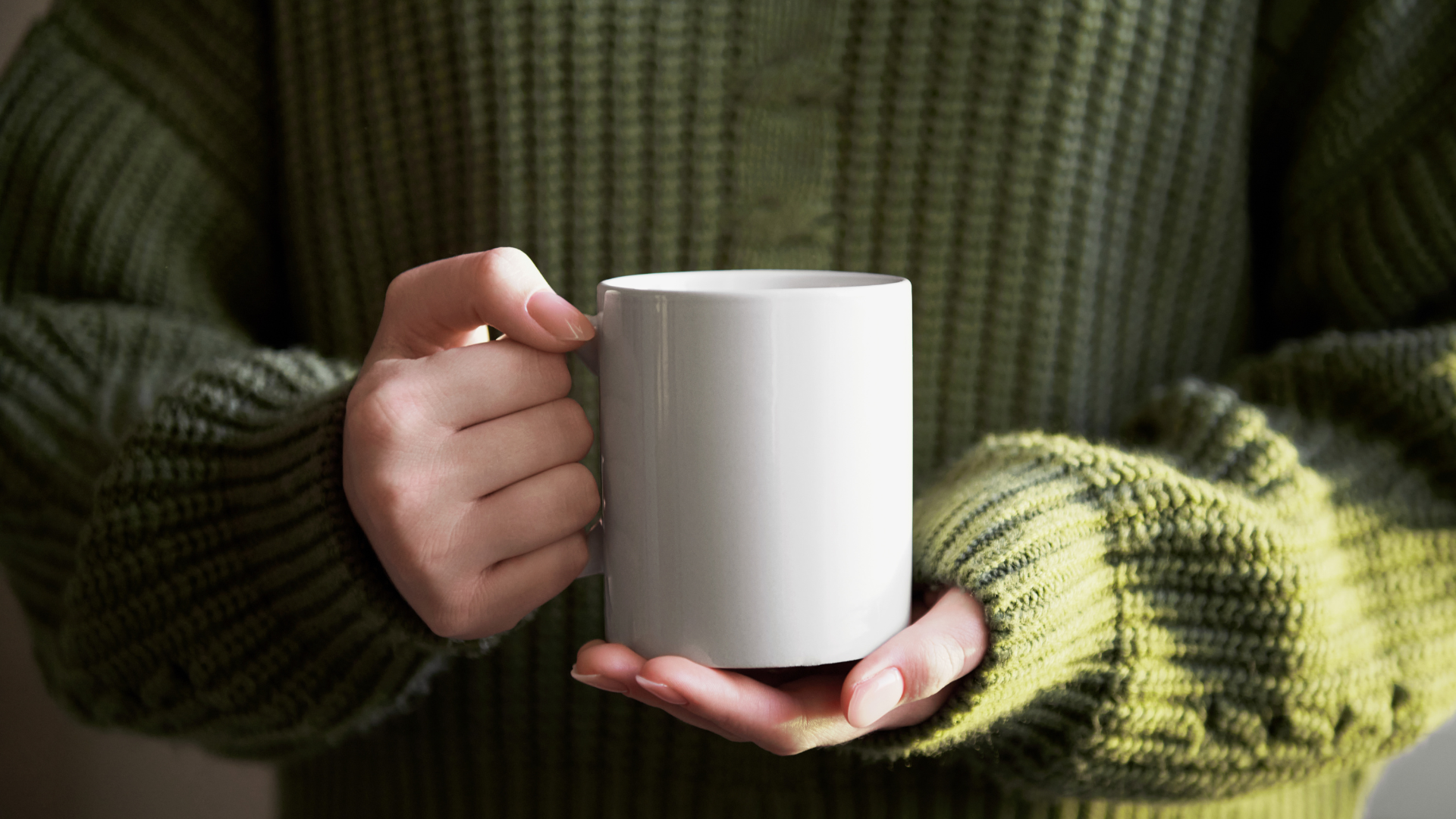 Hands holding a white coffee mug while wearing a dark green sweater.
