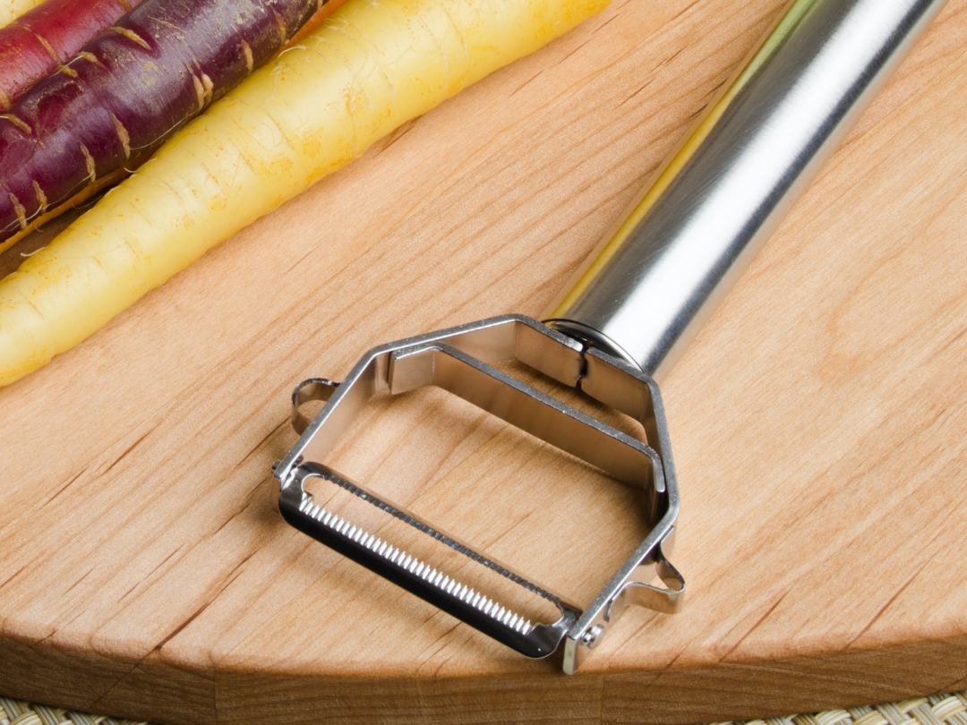 A metal vegetable peeler resting on a wooden cutting board with colorful purple, yellow, and orange carrots lined up nearby.