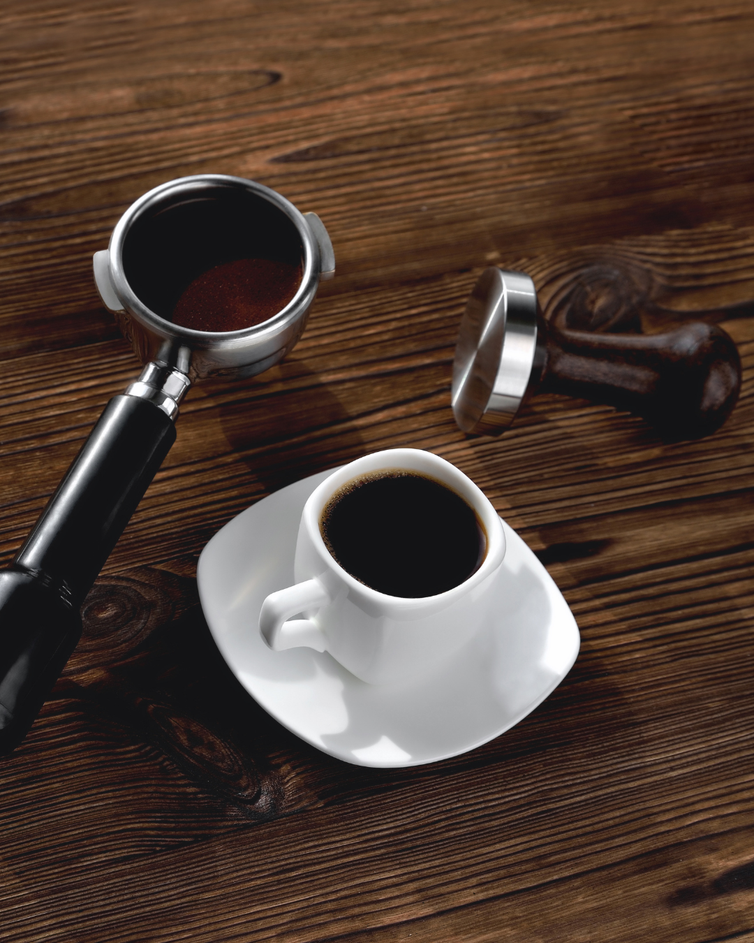 Espresso cup beside a portafilter filled with ground coffee and a coffee tamper on a wooden table.