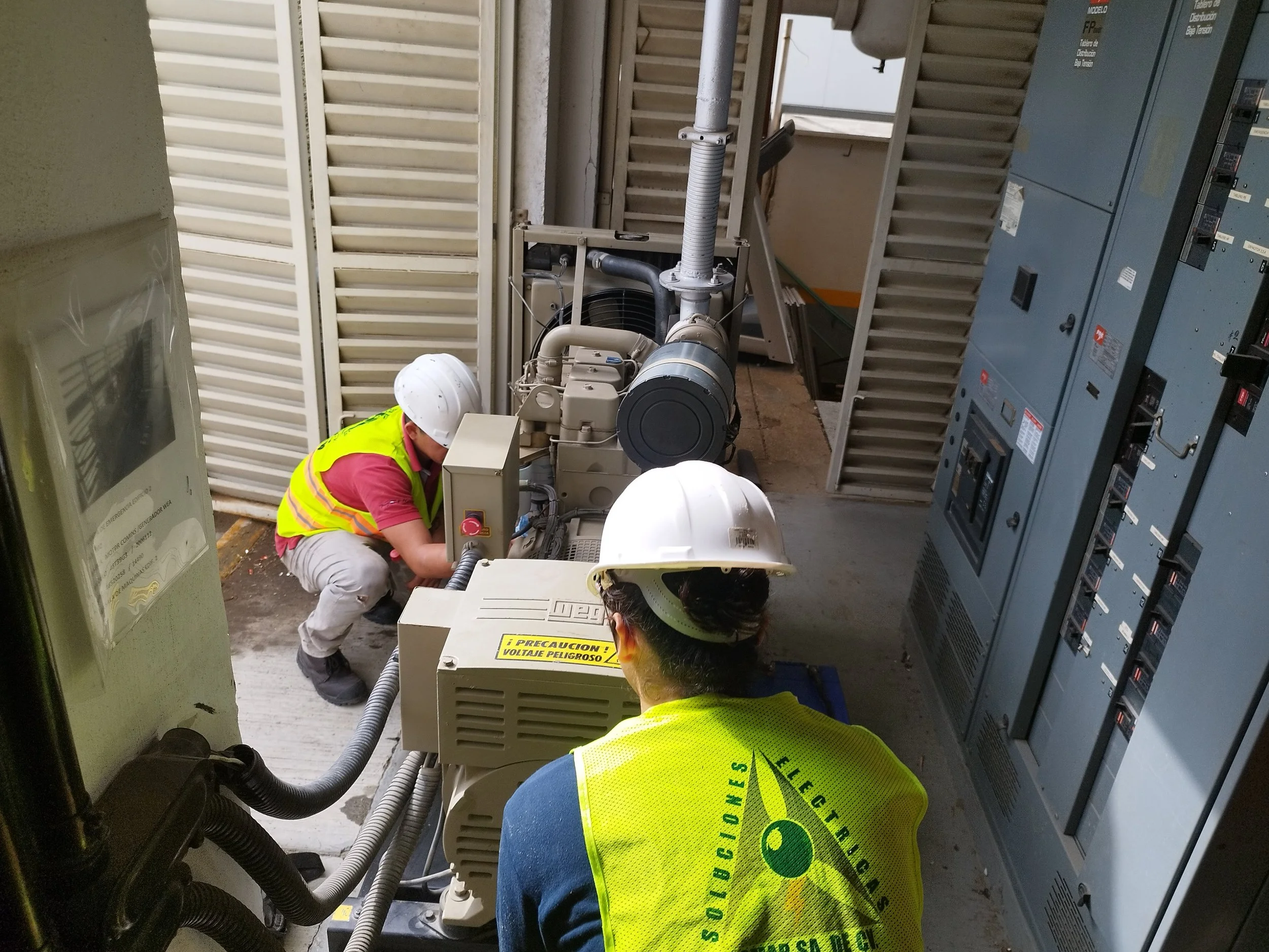 Personal de trabajos de electricidad inspeccionando un equipo eléctrico grande en un lugar industrial, con paneles y cables visibles.