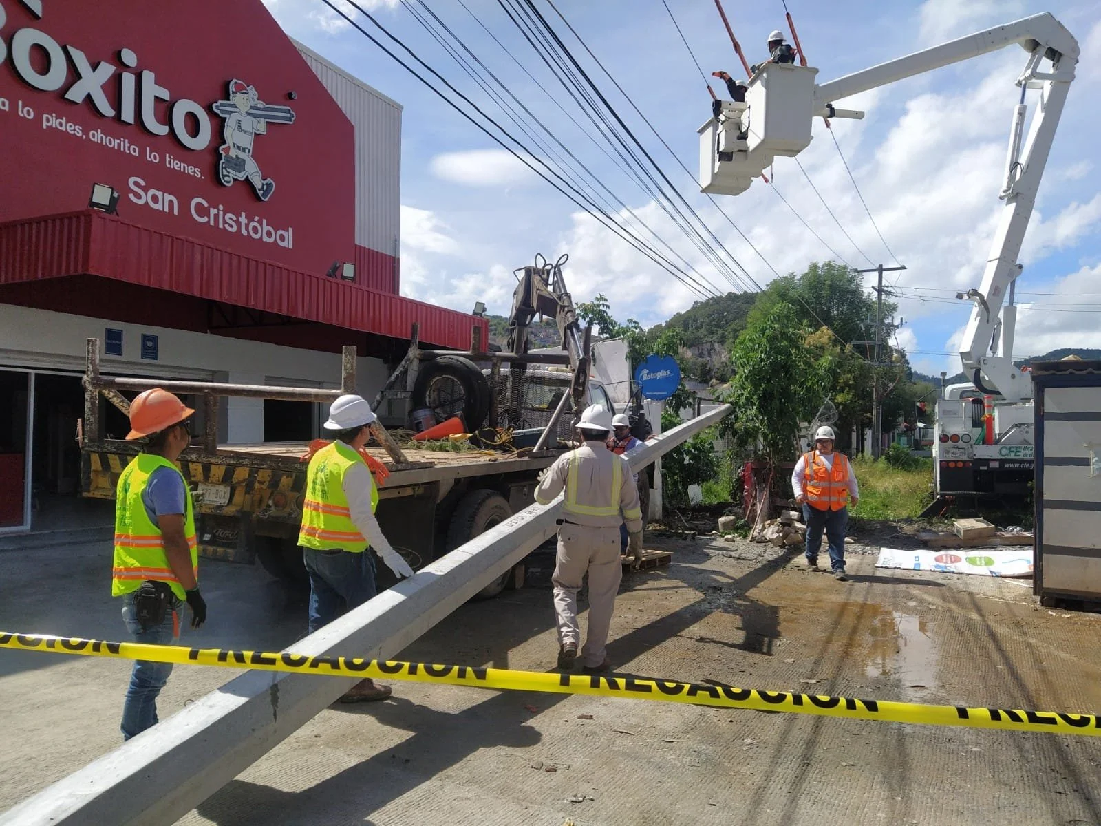 Trabajadores de construcción en una obra cerca de una tienda llamada Oxito, con un camión, equipo y cables eléctricos en un día soleado.