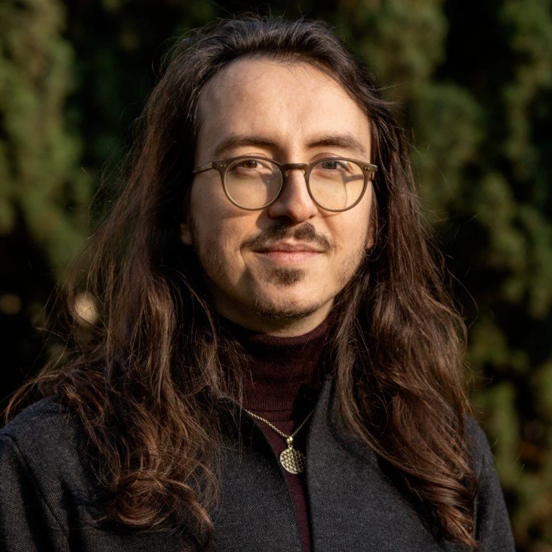 Porträt eines Mannes mit langen braunen Haaren, Brille und dunkler Kleidung, im Freien vor grüner Natur.