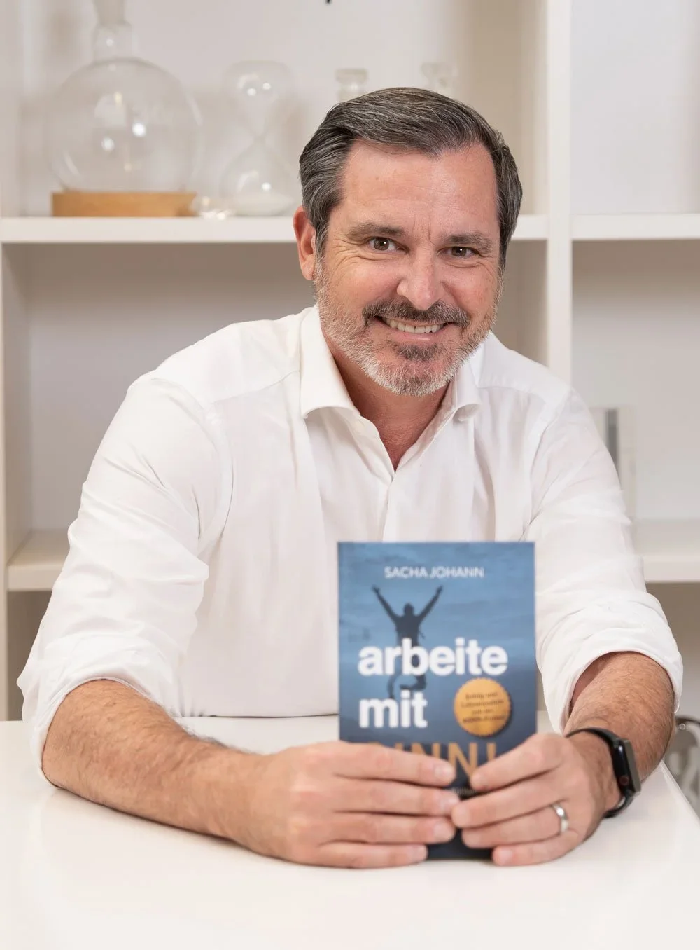 Ein Mann mit grauem Haar und Bart hält ein Buch mit dem Titel »arbeite mit MIN«, sitzend an einem weißen Tisch in einem Raum mit weißen Regalen im Hintergrund.