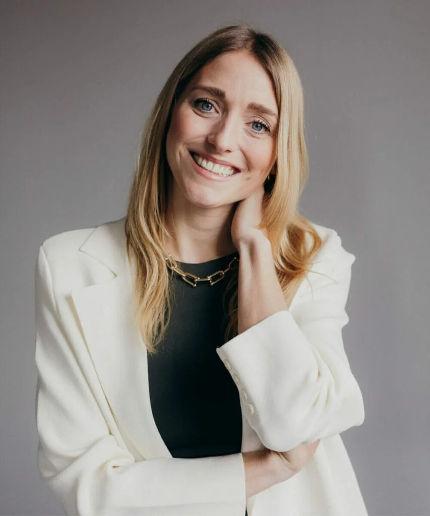 Eine lachende Frau mit blonden, gewellten Haaren, weißem Blazer, schwarzem Oberteil und goldener Kette poses auf einem grauen Hintergrund.