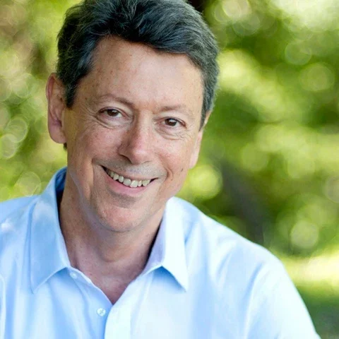 Ein lächelnder Mann mit dunklem Haar, trägt ein blaues Hemd, im Hintergrund sind grüne Bäume und Sonnenlicht.