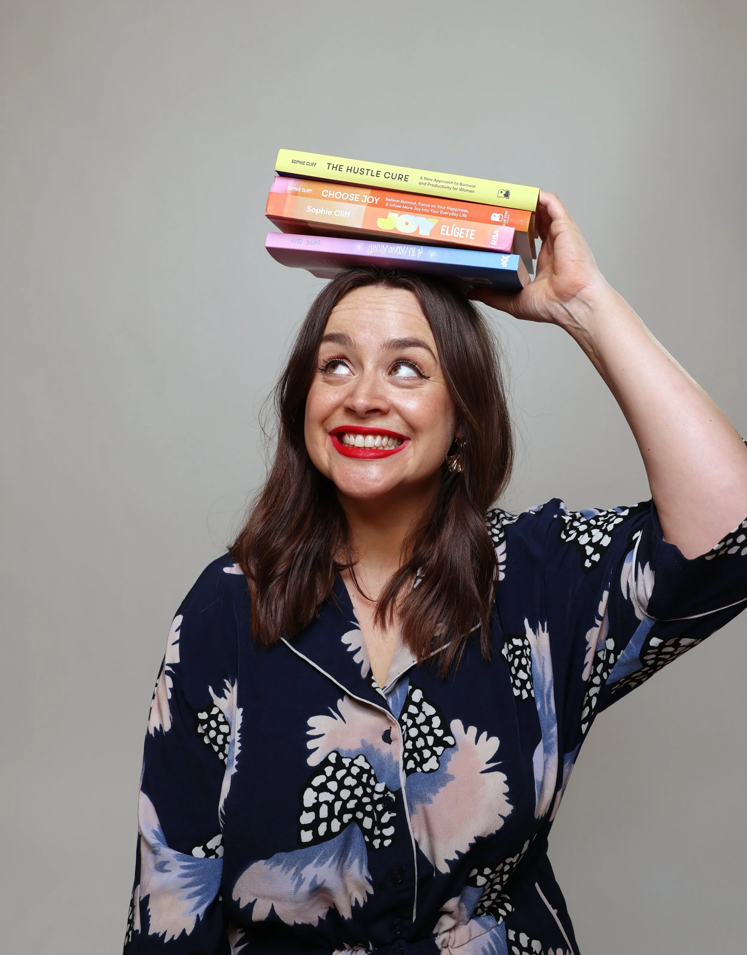Eine lachende Frau mit braunem Haar trägt ein blau-weiß gemustertes Kleid und hält drei Bücher auf dem Kopf.