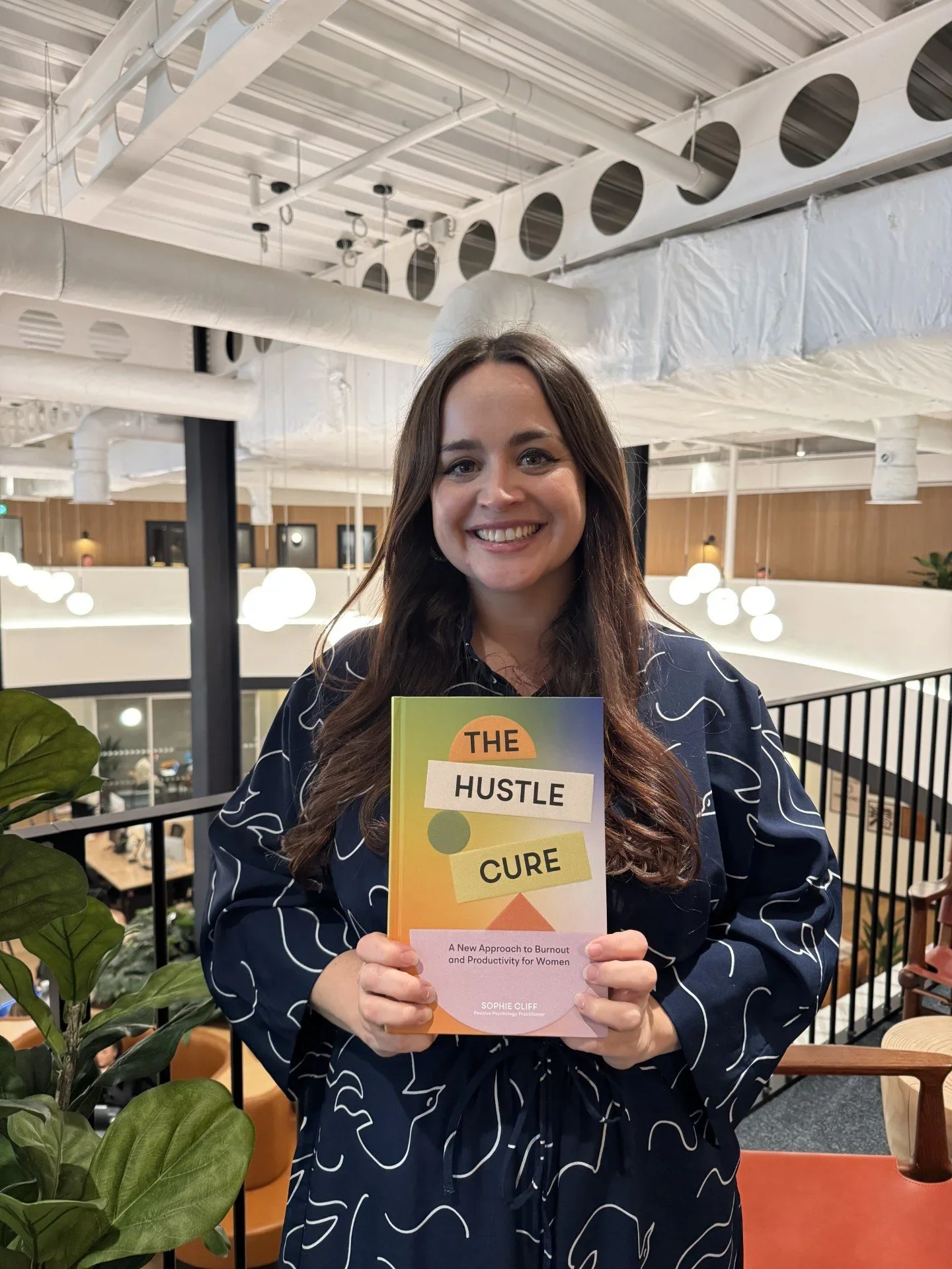 Eine lächelnde Frau hält ein Buch mit dem Titel „The Hustle Cure“. Sie befindet sich in einem modernen Innenraum mit weißer Decke, runden weißen Lampen und Holzpaneelen an der Wand. Im Vordergrund sind grüne Pflanzen sichtbar.