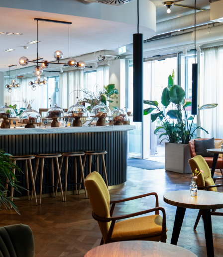 The Studio Kaffee von Ringier - Innenansicht eines modernen Cafés oder Restaurants mit Tischen, Stühlen und großen Pflanzen, viel natürliches Licht durch große Fenster.