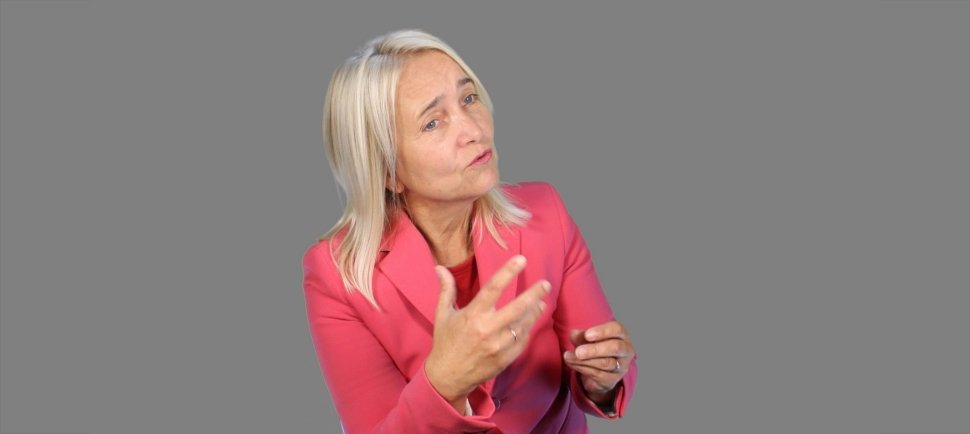 Ältere Frau mit blondem Haar in pinkem Blazer, die skeptisch blickt und die Hand hebt, vor grauem Hintergrund.