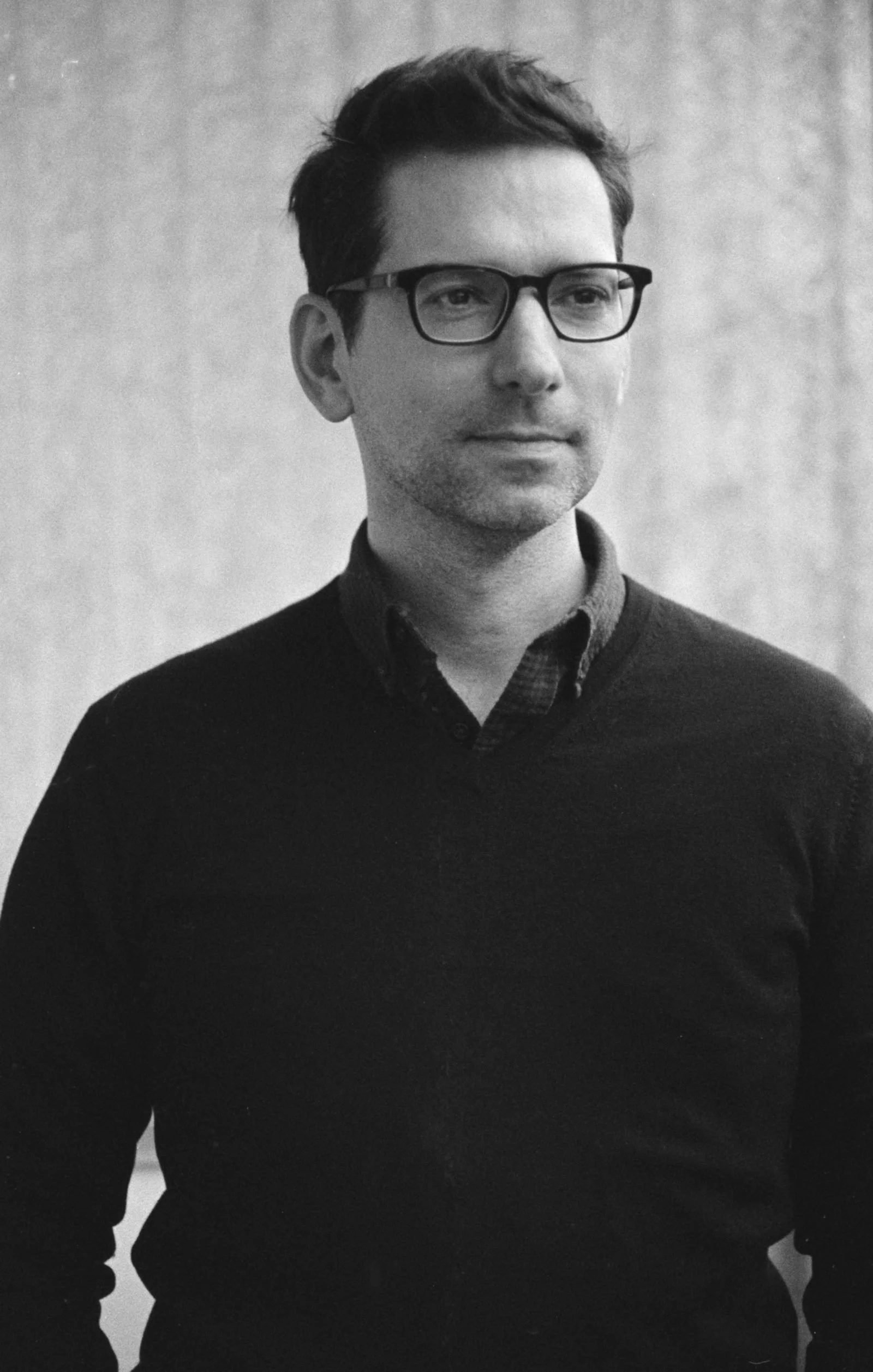 Schwarz-Weiß-Porträt eines Mannes mit dunklem Haar, Brille und dunklem Pullover, vor hellem Hintergrund.