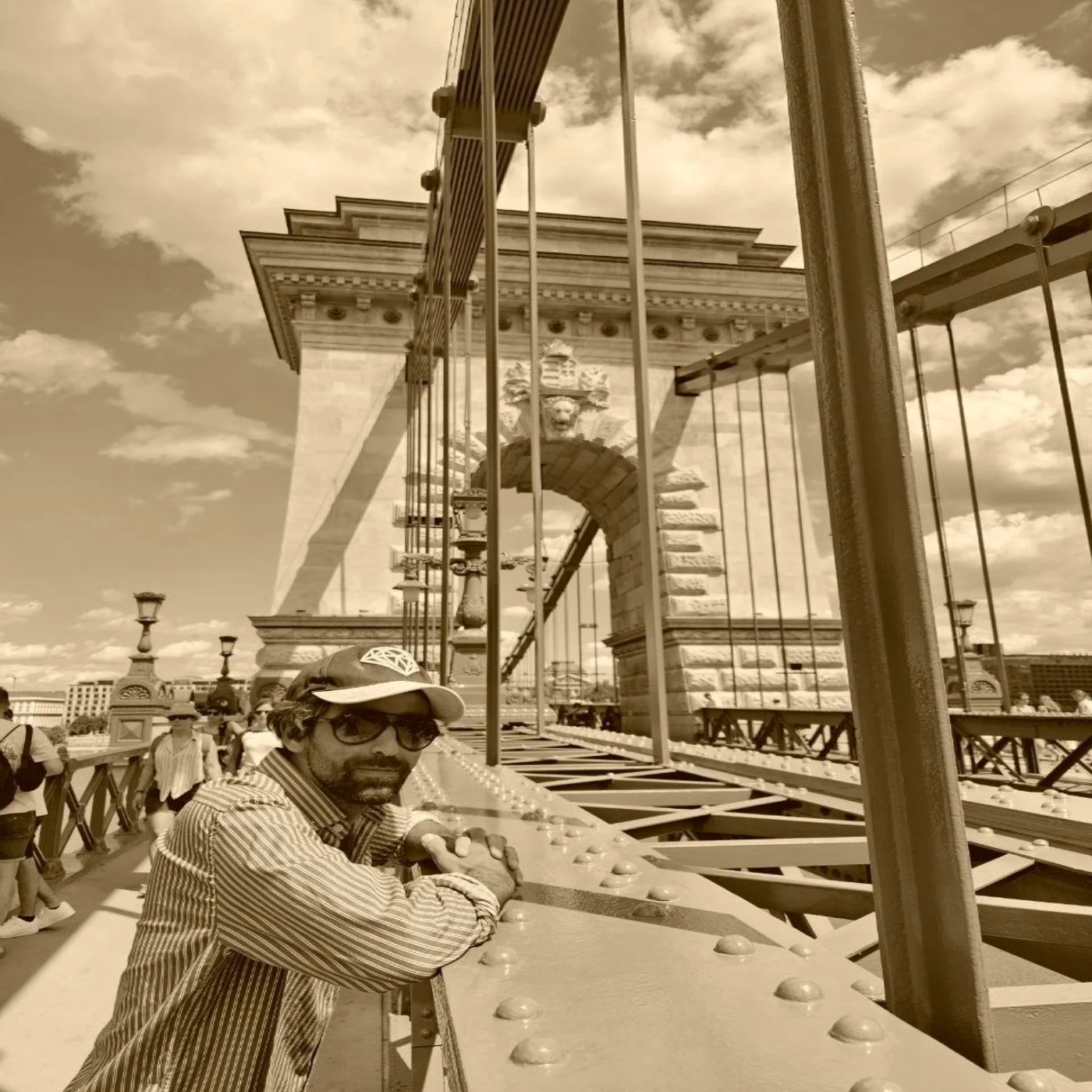 A man wearing sunglasses and a cap stands on a bridge or scaffolding with metal beams, in front of a historic stone arch with a lion sculpture, part of a large stone structure, possibly a historic monument or bridge, under a partly cloudy sky with a cityscape in the background.