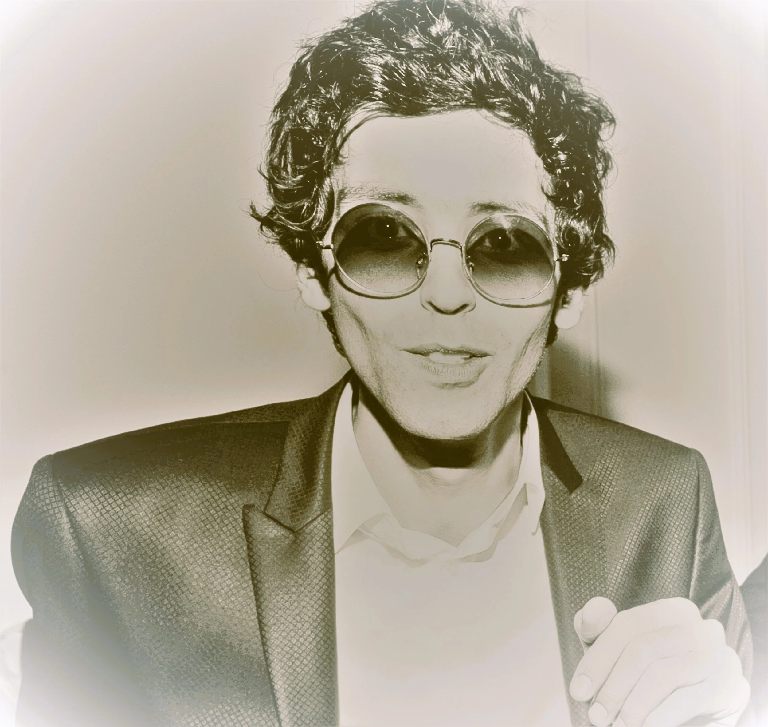 A person with curly hair wearing tinted round sunglasses and a checkered blazer. They are holding a coffee cup and have a serious expression.
