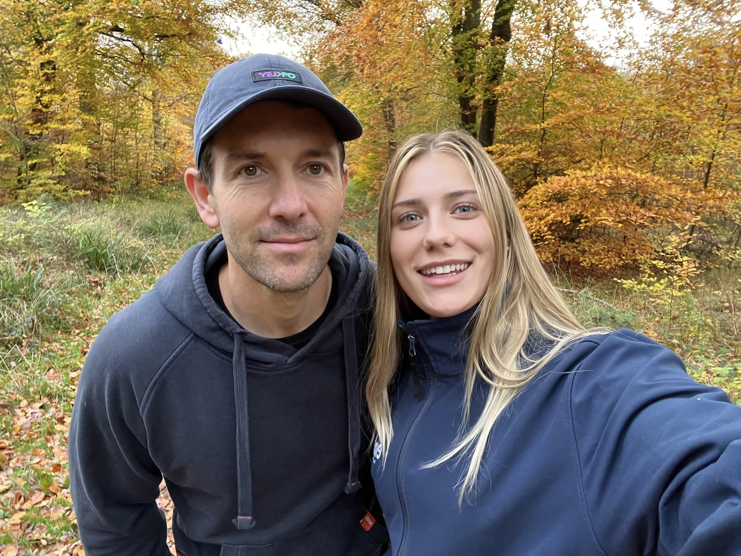 A man and a woman taking a selfie outdoors in an autumnal forest, with colorful fall foliage and leaves on the ground.