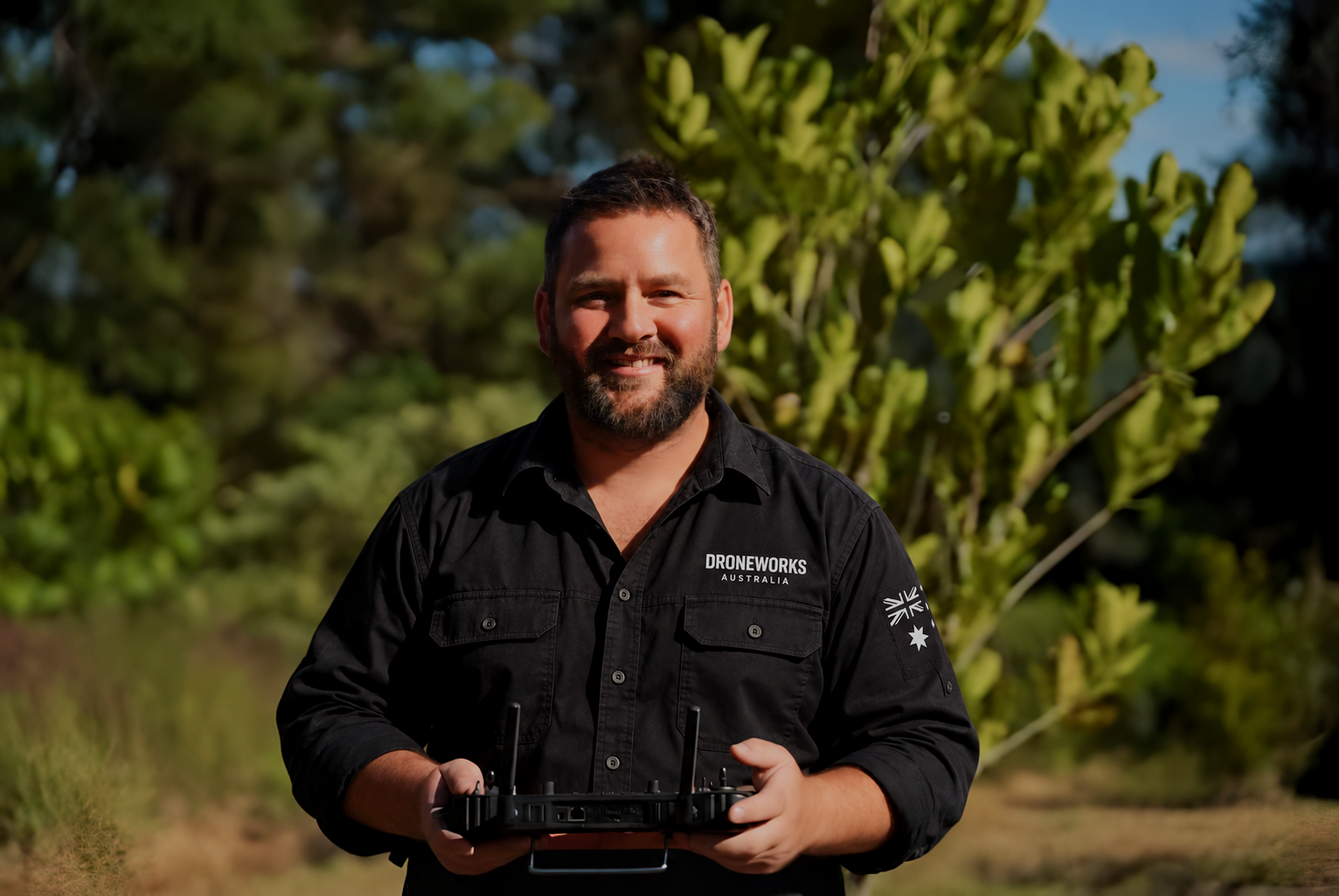 A man with a beard and short hair smiling outdoors, holding a drone controller, wearing a black shirt with 'DRONEWORKS AUSTRALIA' logo and a patch of the Australian flag.