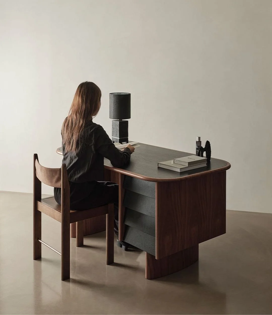 A woman with long brown hair sitting at a wooden desk, looking at her phone, with a black table lamp, a stack of books, and some small objects on the desk, against a plain light-colored wall.