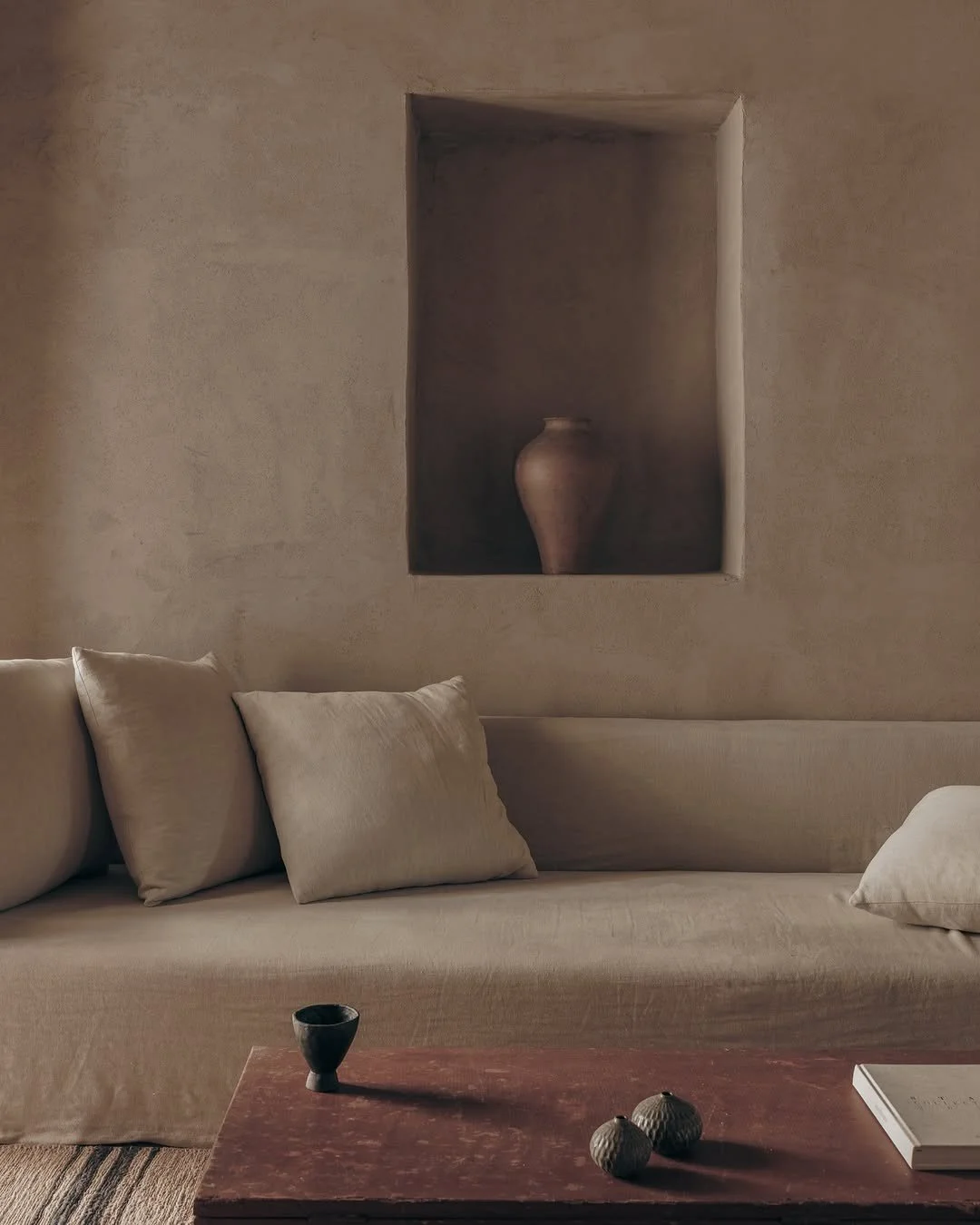 A beige sofa with three matching pillows, a red wooden coffee table with a black cup, two decorative vases, and a white book. There is a square inset on the wall with a large clay vase inside.