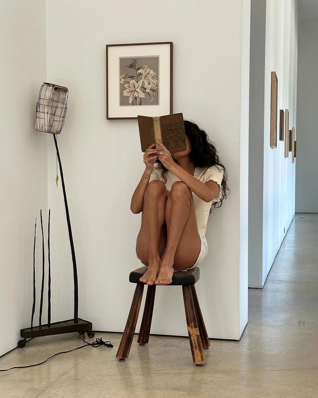 A woman with long curly hair sitting on a wooden stool in a white room, reading a book with her knees pulled up to her chest. There is a framed floral picture on the wall behind her, and a tall floor lamp with a patterned shade to her left.