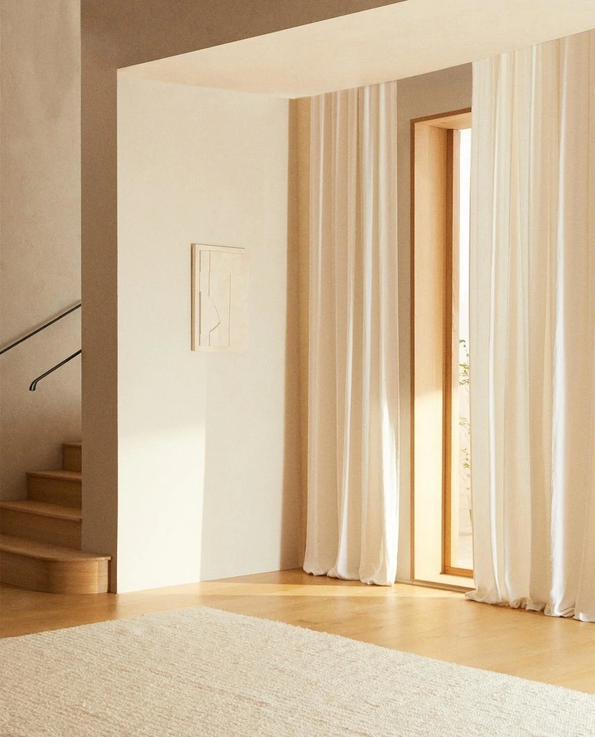 Sunlit interior space with a wooden framed window and beige curtains, staircase with wooden steps, and cream-colored walls.