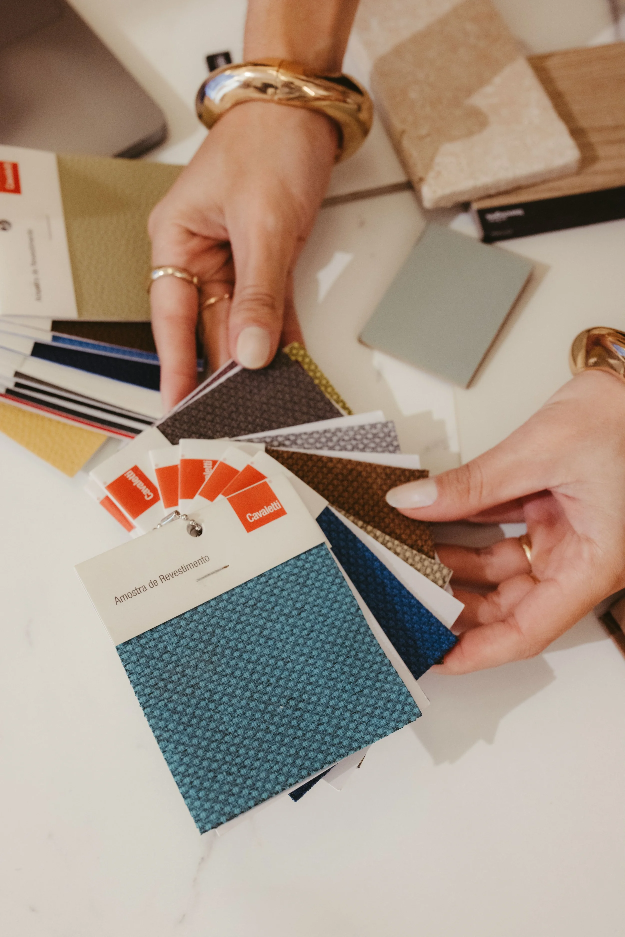 Person examining fabric samples and paint color swatches on a white table, wearing a gold bracelet and rings.