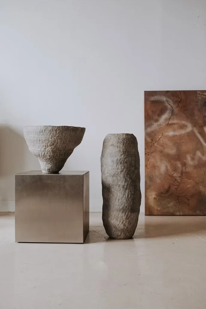 Two stone sculptures, one on a metallic pedestal and one standing on the floor, with a painting in the background.