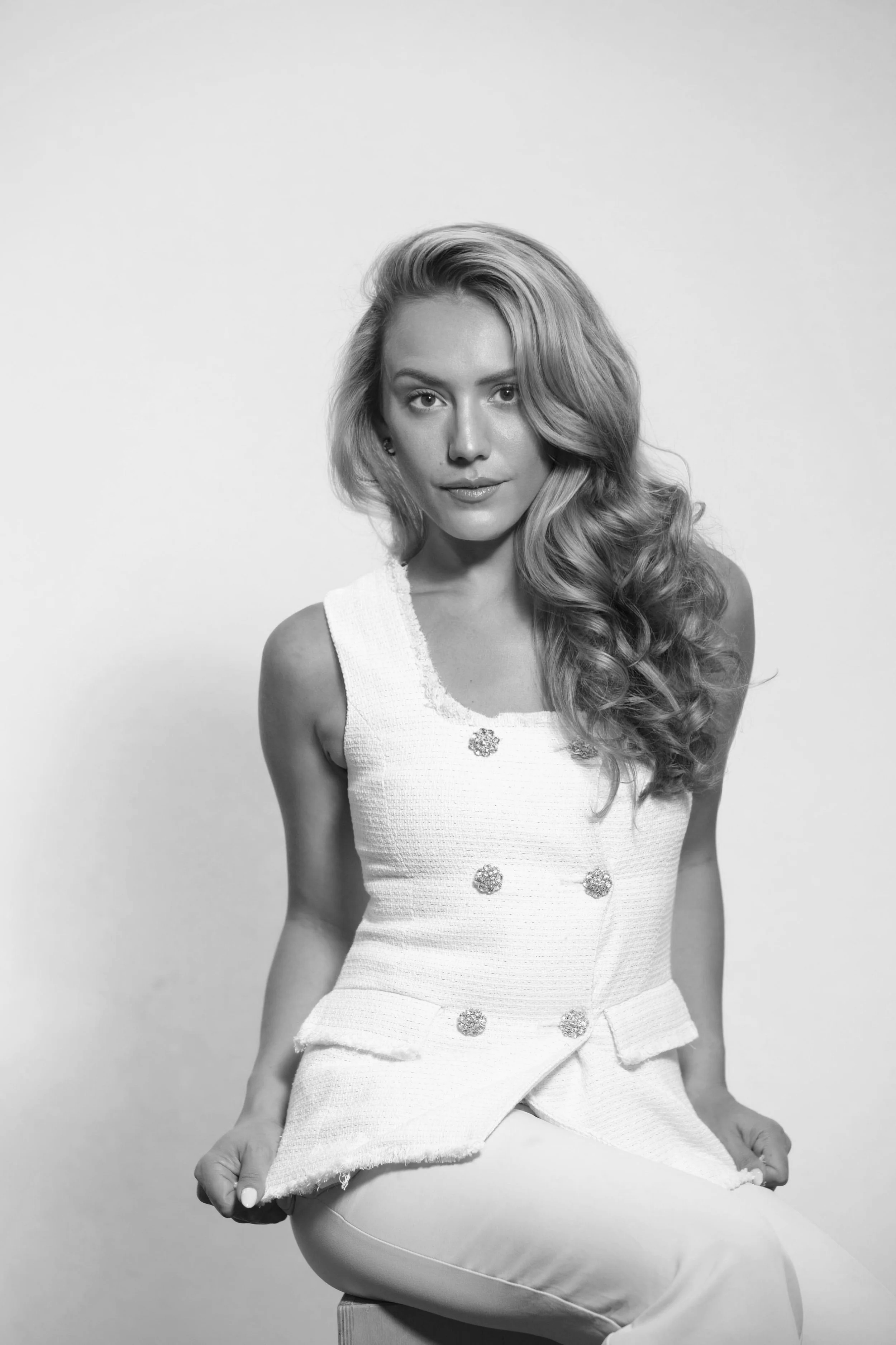 Black and white photo of a young woman with long wavy hair sitting on a stool, wearing a sleeveless dress with floral buttons, posing with one knee up and hands resting on her knee against a plain background.