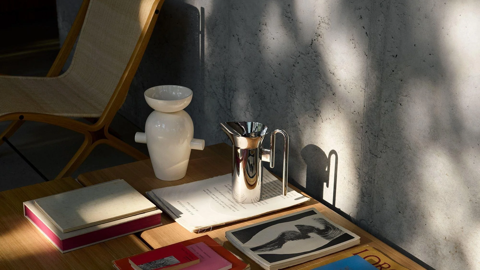 A tabletop with books, a metallic pitcher, a white ceramic vase, a bowl, and papers, placed against a textured concrete wall, with sunlight creating shadows.