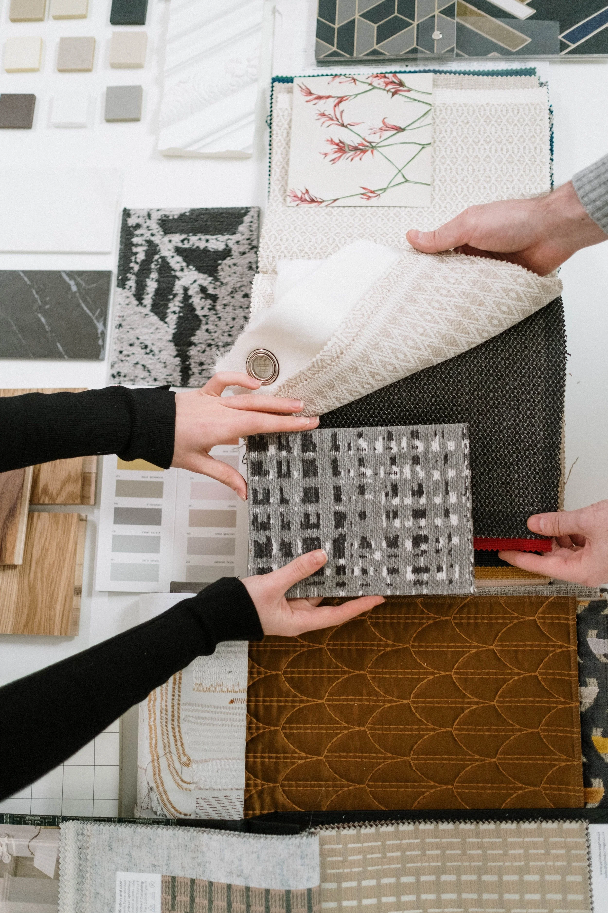 Various fabric swatches and samples of different patterns, colors, and textures displayed on a table, some being held by hands.