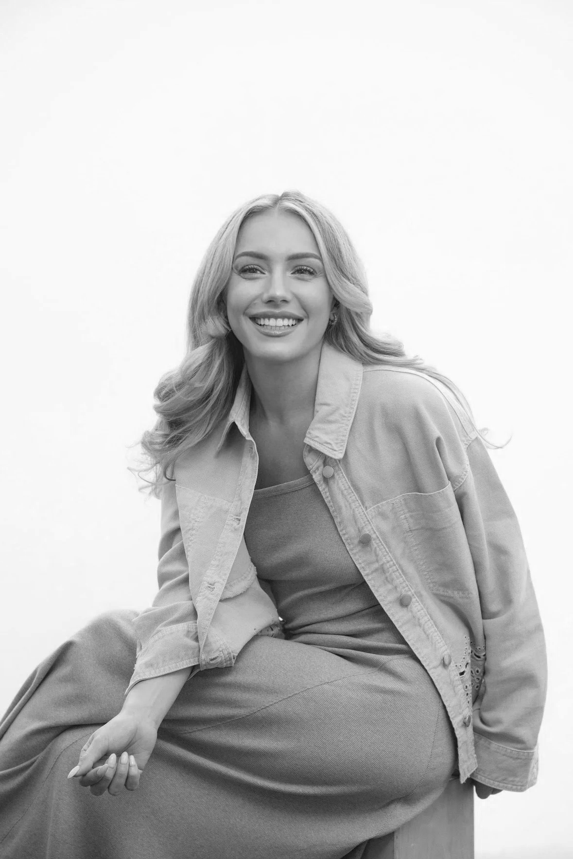 Black and white photo of a young woman with long wavy hair smiling, wearing a casual denim jacket and sitting on a large rounded object, against a plain white background.