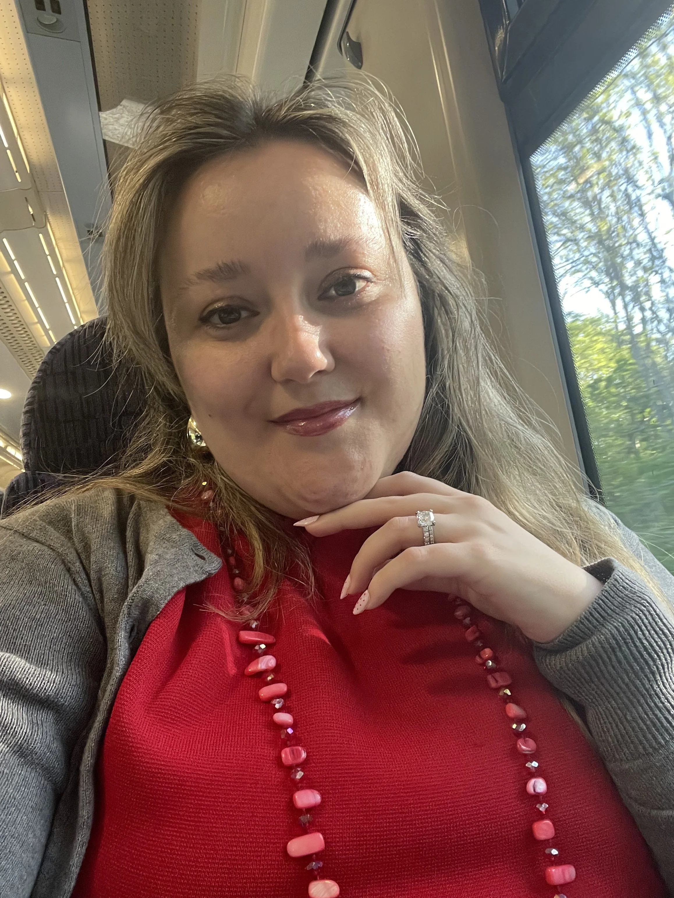A woman with shoulder-length hair, wearing a red top, gray jacket, and beaded jewelry, is sitting in a train with a window showing trees outside. She has a ring on her finger and is smiling at the camera.