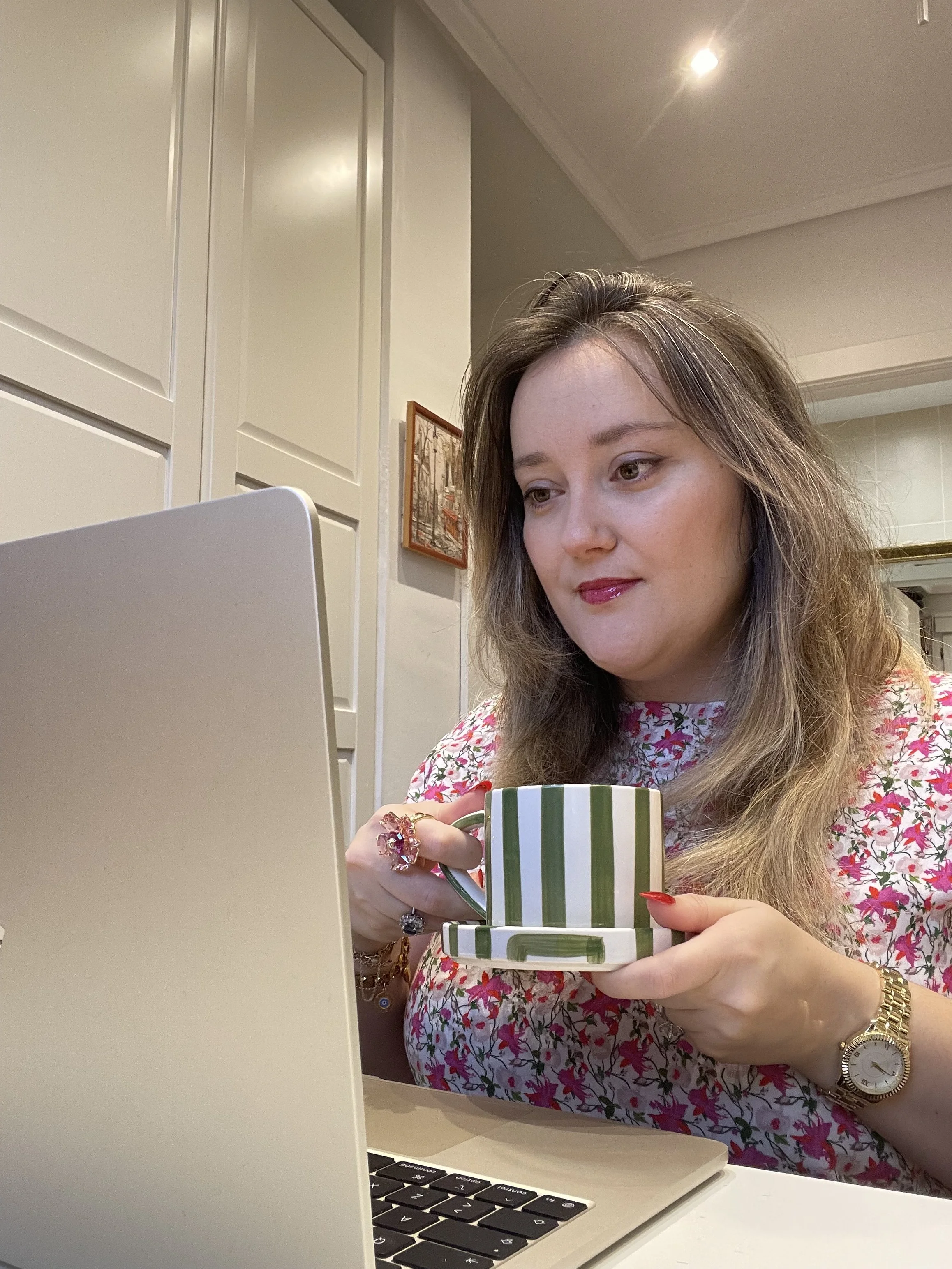 A woman with long, wavy hair, wearing jewelry and a floral dress, is using a laptop at a desk. She is holding a green and white striped mug in her left hand and is looking at the screen. A keyboard is visible in the foreground, and there is a framed picture on the wall behind her.